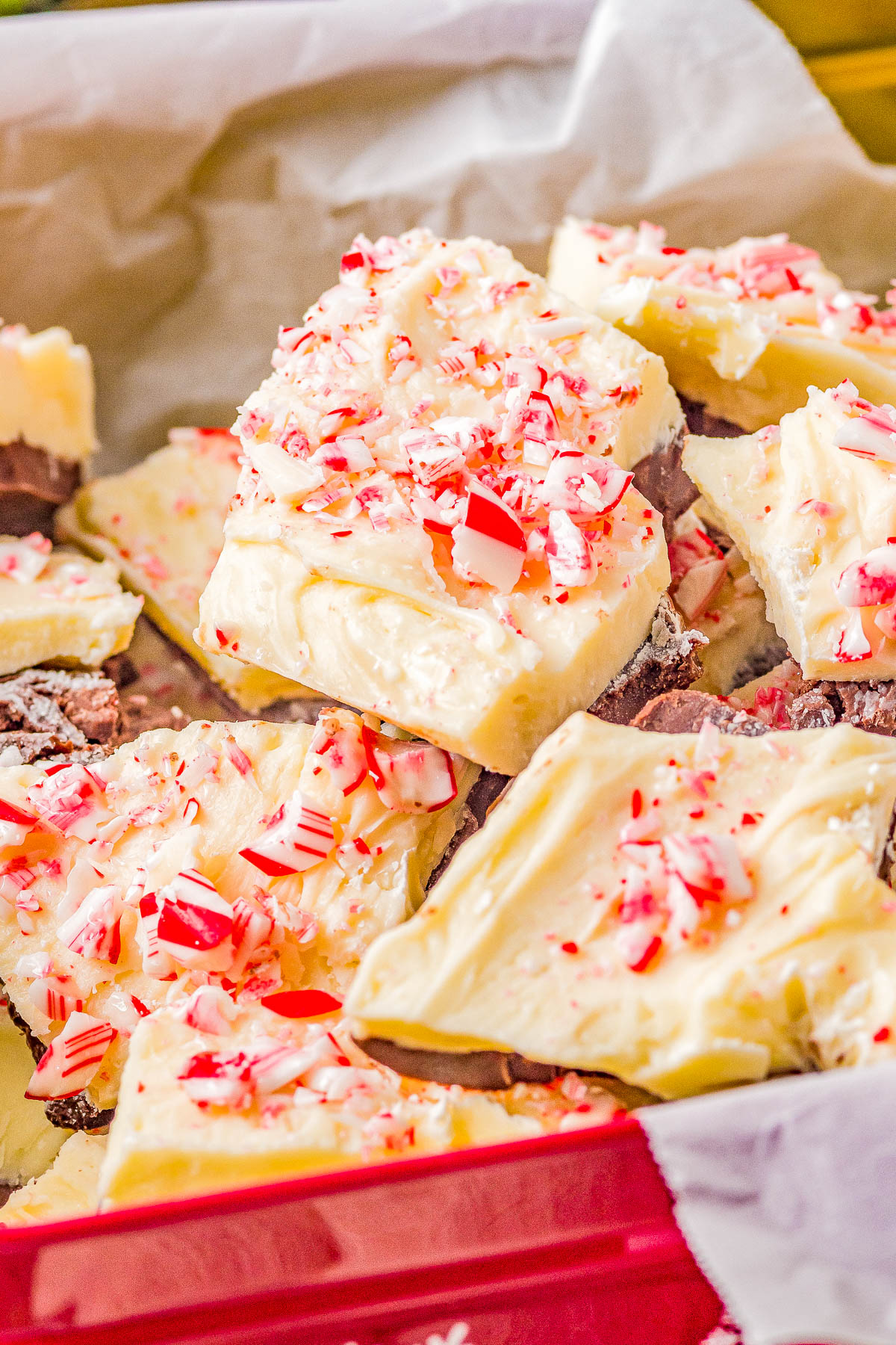 Pieces of white chocolate peppermint bark with crushed candy canes on top, stacked in a red tray lined with parchment paper.