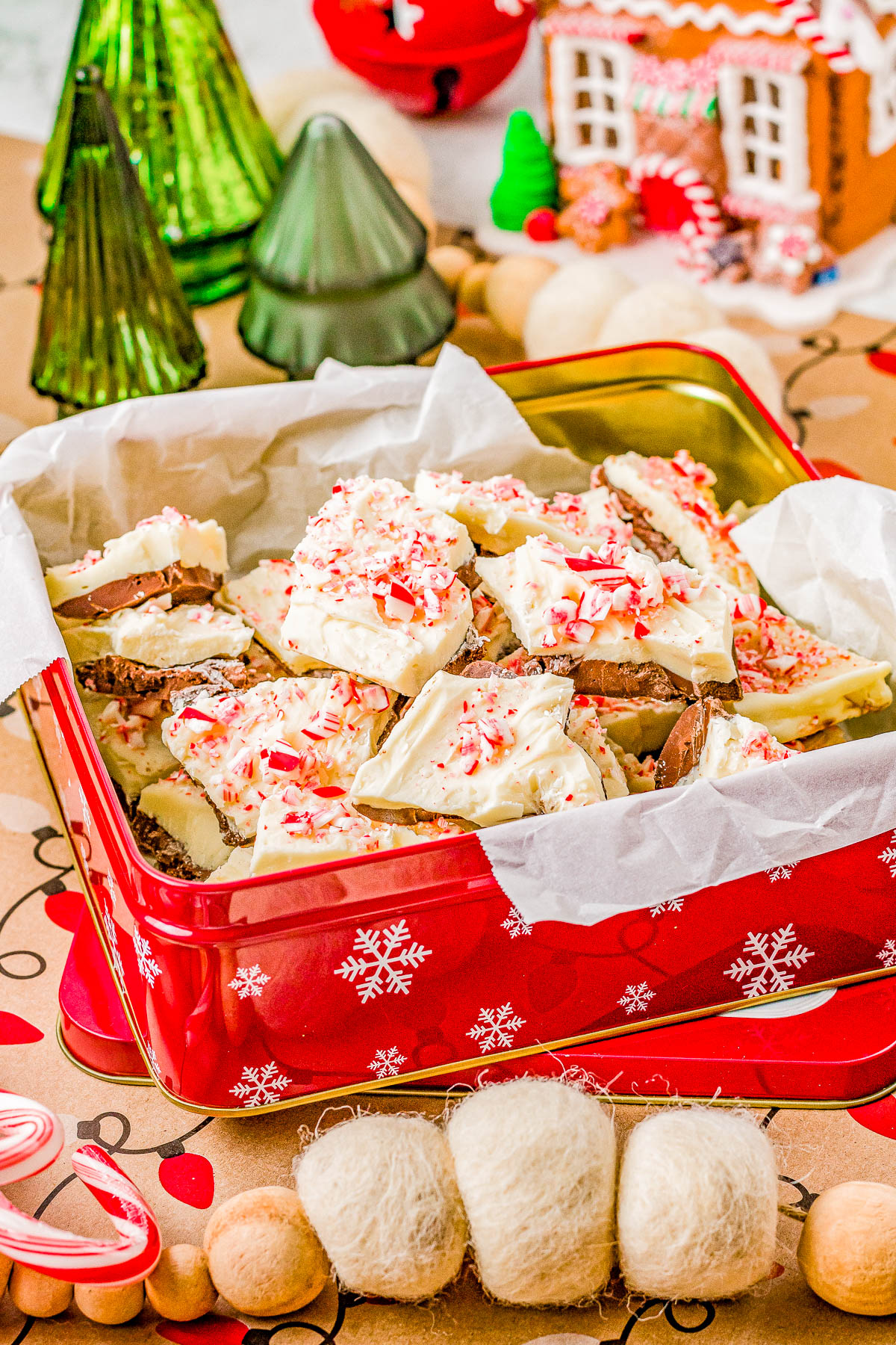 A festive tin filled with peppermint bark on parchment paper. The bark is topped with crushed candy canes. Decorated Christmas trees and a gingerbread house are in the background.