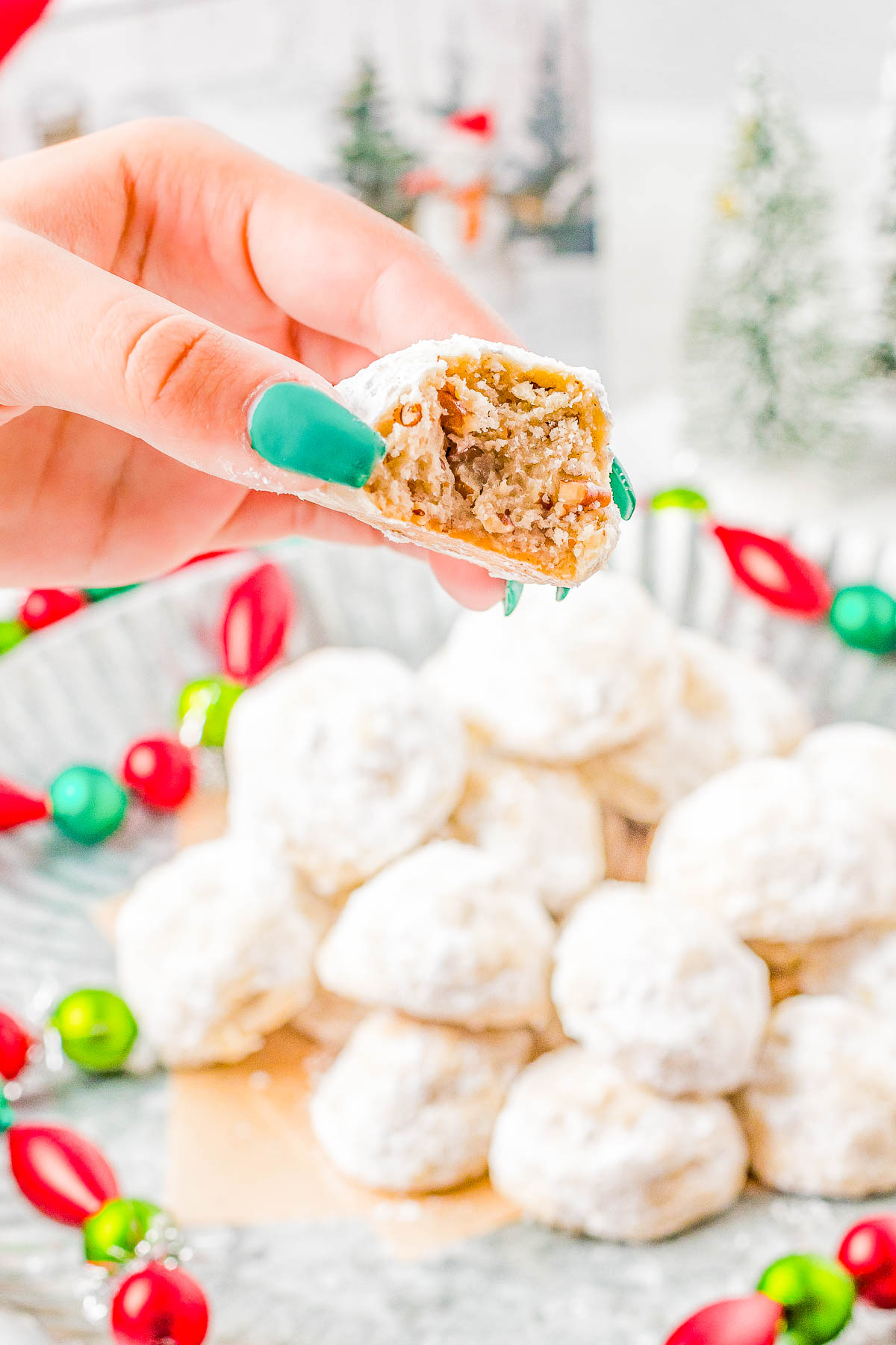 A hand with teal nail polish holds a bitten snowball cookie above a plate of similar cookies. The cookies are topped with powdered sugar, and decorative ornaments are in the background.