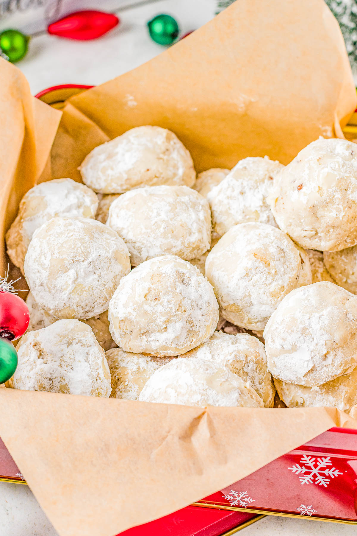 A tray of round, powdered sugar-coated cookies, arranged on brown parchment paper. Festive ornaments are partially visible in the background.