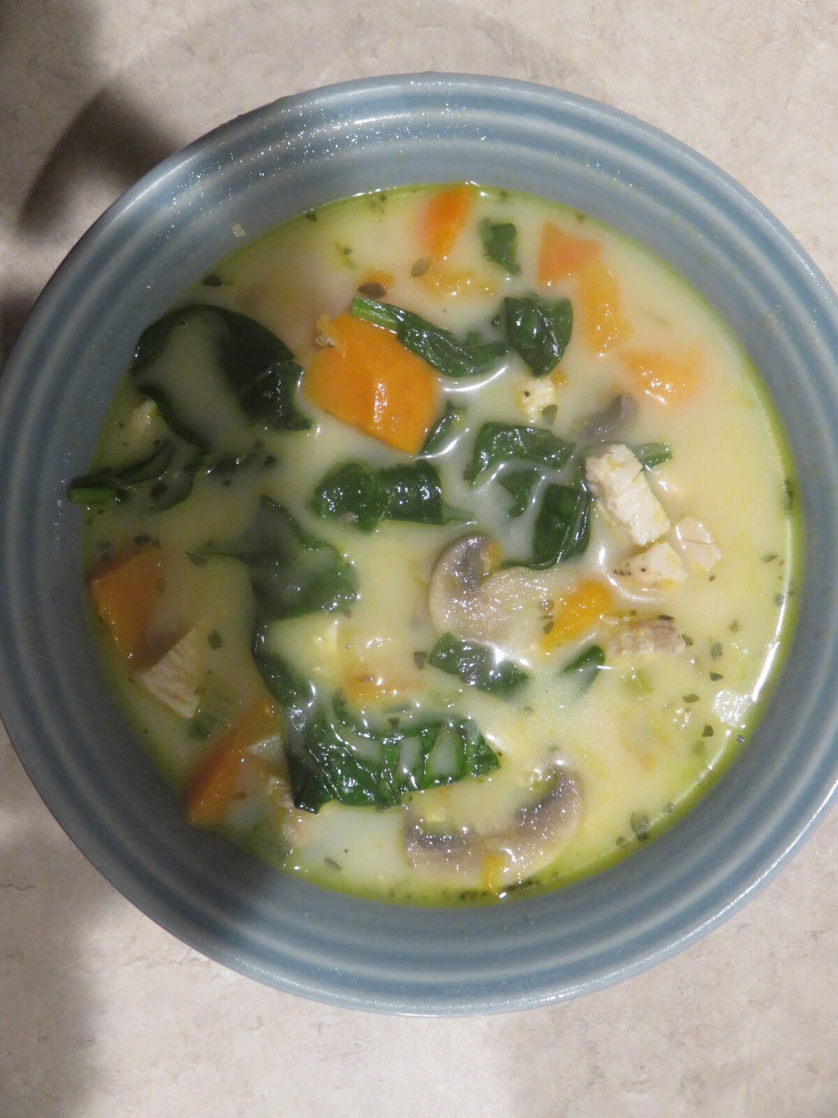 A blue bowl filled with creamy soup containing spinach, orange bell peppers, mushrooms, and chunks of chicken.