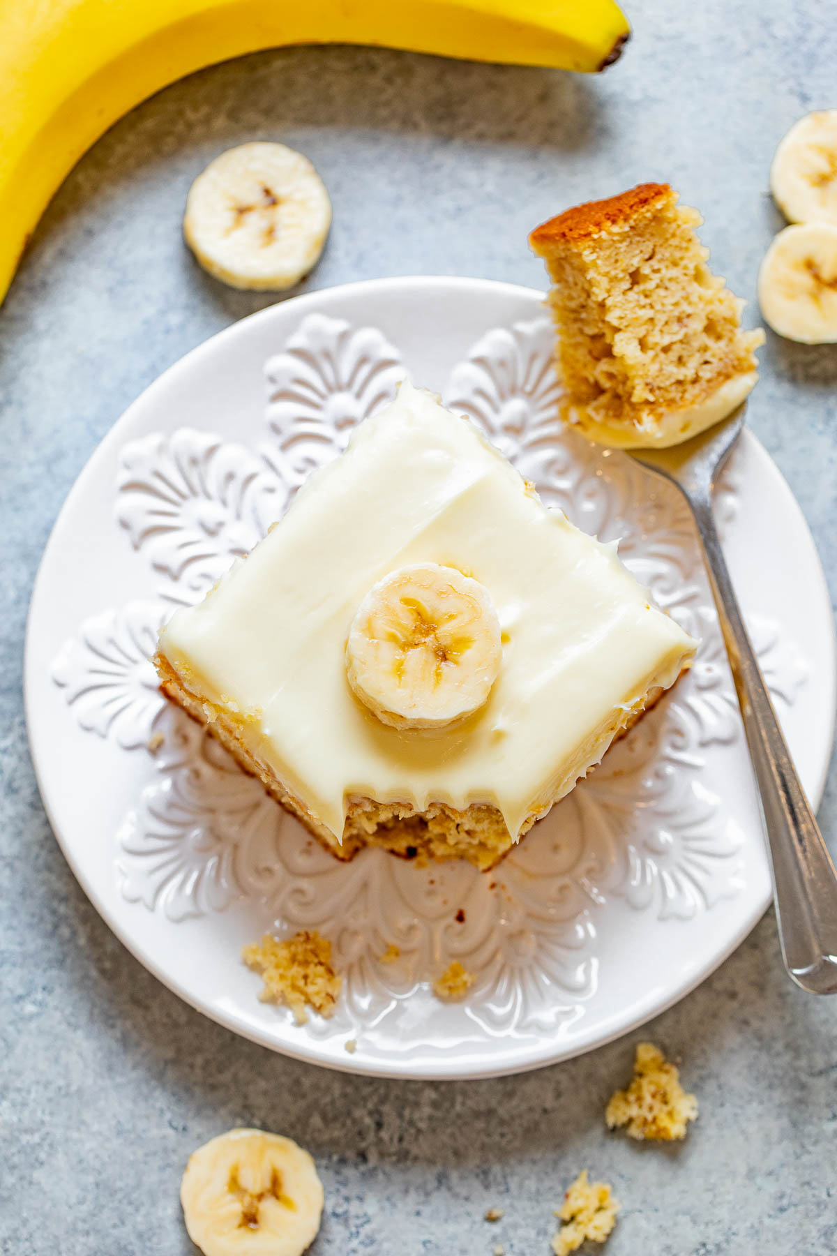 A square piece of banana cake with cream cheese frosting on a decorative white plate. A banana slice tops the cake, and a fork with cake crumbs is beside it. Several banana slices are scattered around.