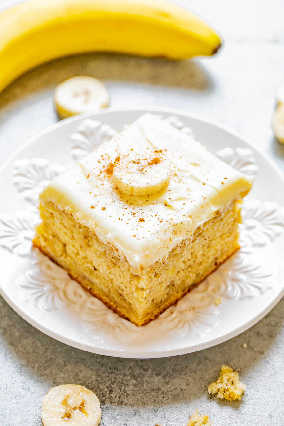 A slice of banana cake with white frosting and a banana slice on top, served on a white plate. A whole banana and banana slices are in the background.