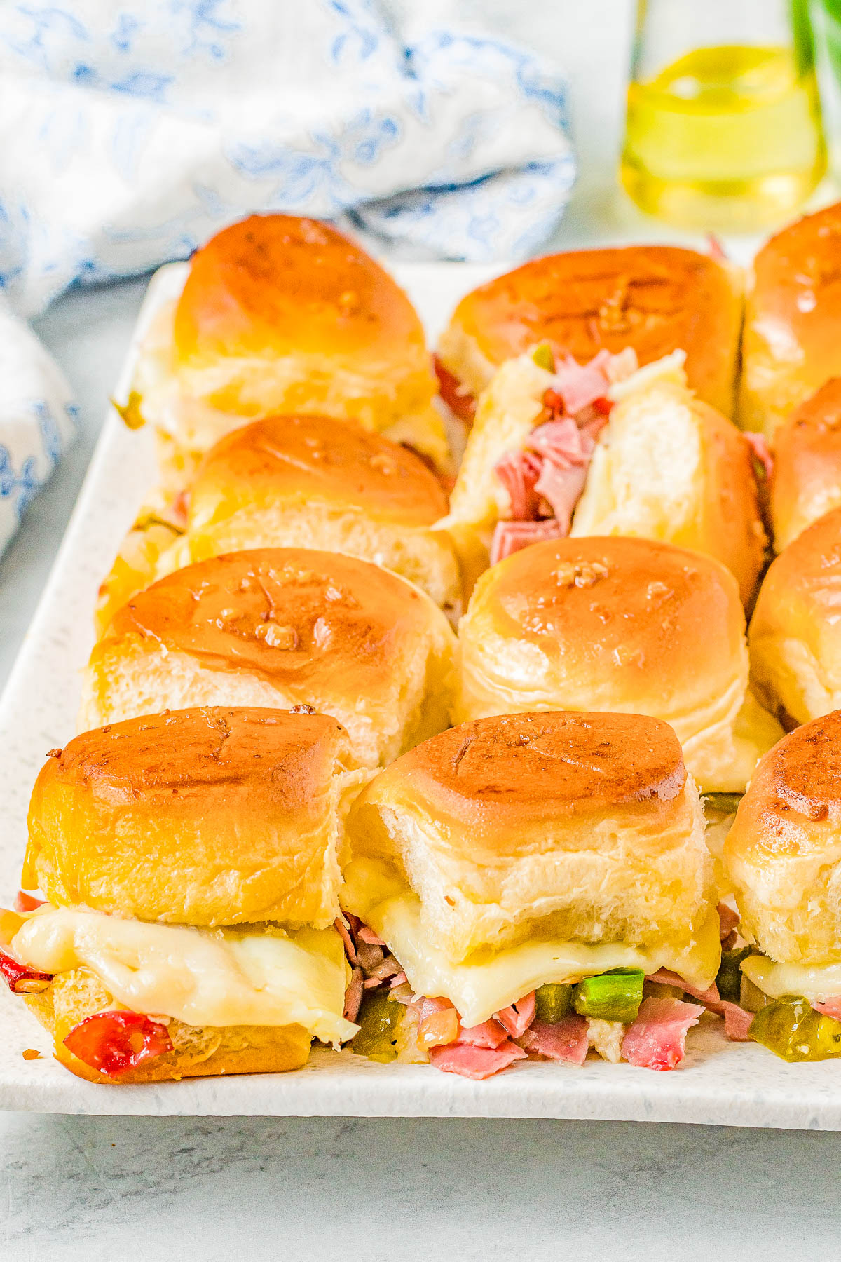 Platter with ham and cheese sliders on buns, topped with melted cheese and garnished with chopped peppers, on a white plate.