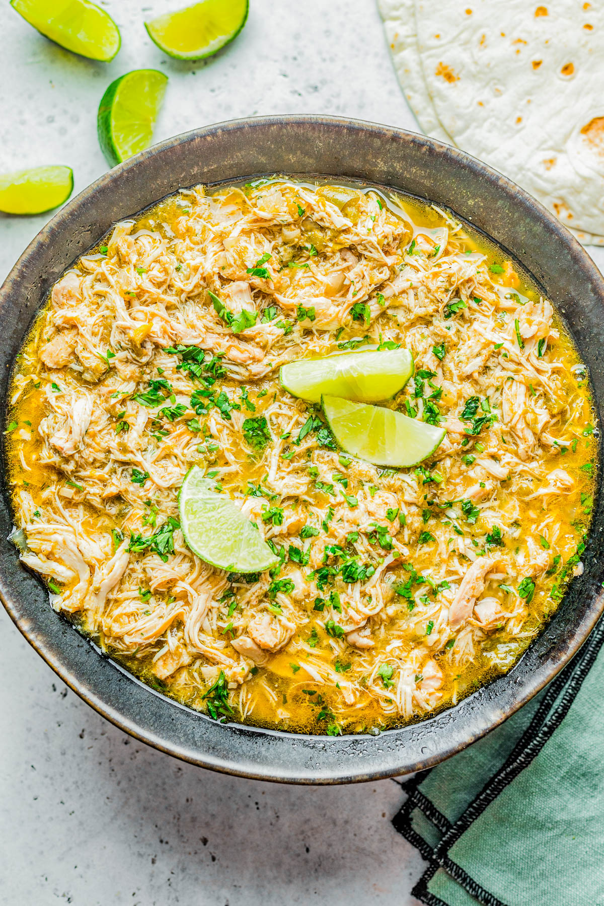 A bowl of shredded chicken in a green sauce, garnished with lime wedges and cilantro, next to lime slices and a tortilla on a white surface.