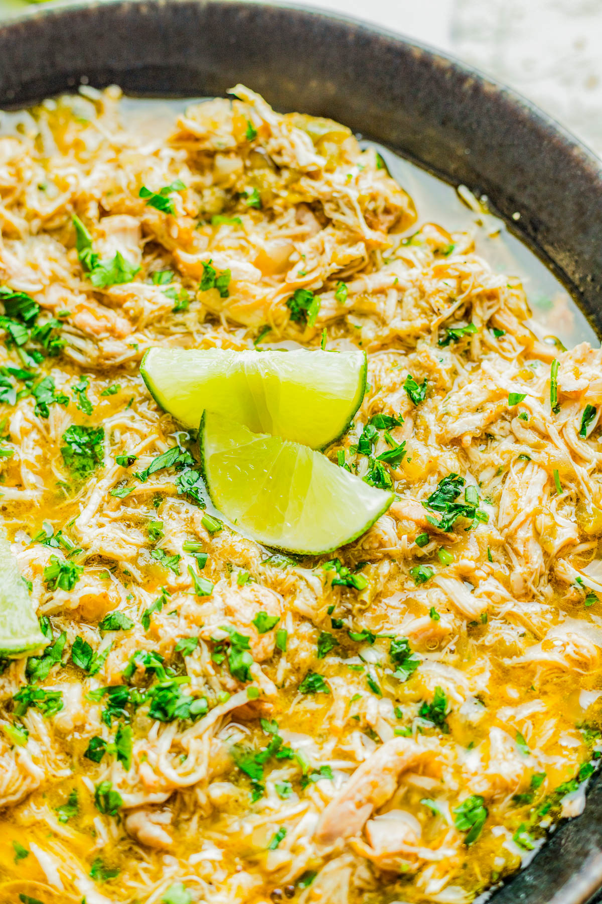 A skillet filled with shredded chicken in a creamy sauce, garnished with chopped herbs and lime wedges on top.