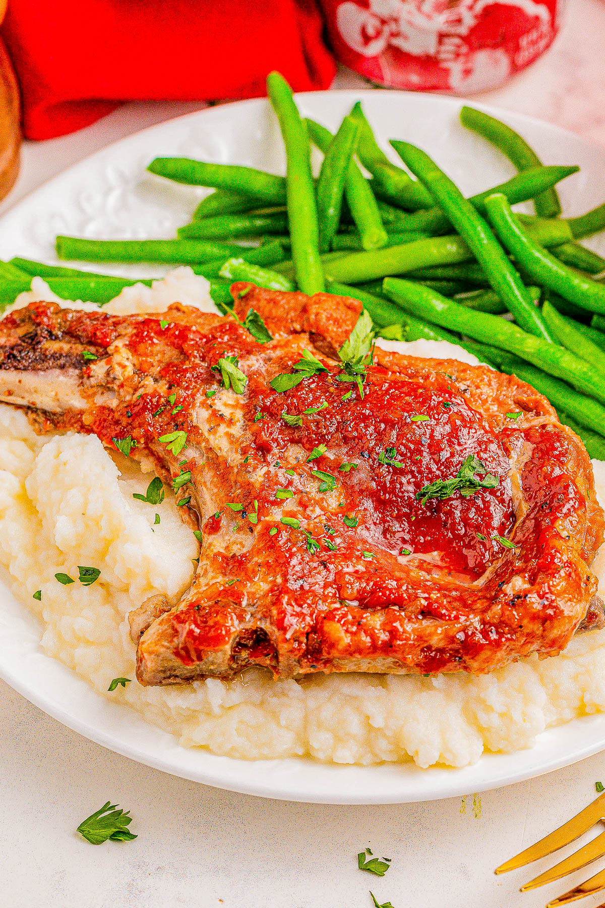 A cooked pork chop with tomato sauce on a bed of mashed potatoes, garnished with herbs, served alongside green beans on a white plate.