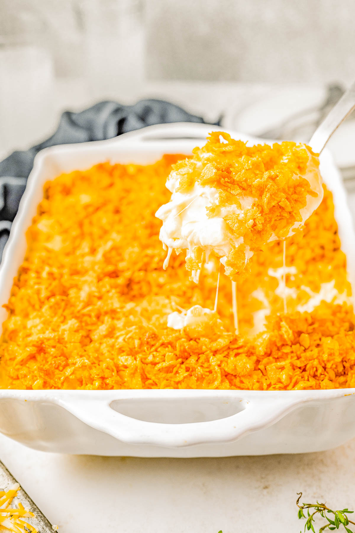 A white baking dish is filled with a baked casserole topped with a golden, crunchy layer. A serving is being lifted with a spoon, showing a creamy interior.