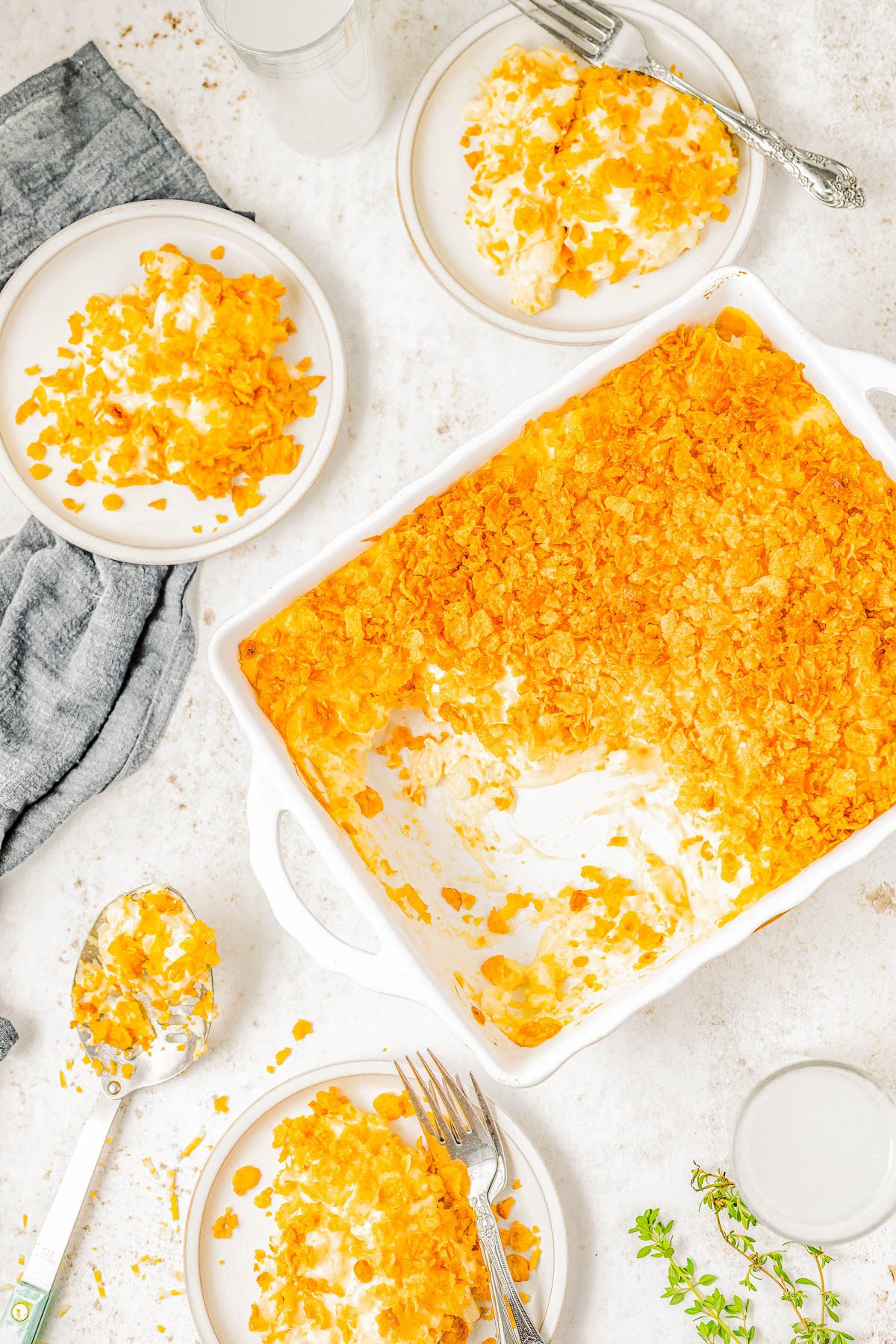 A baking dish filled with a cornflake-topped casserole, partially served onto three plates with forks. A metal serving spoon and a grey cloth are on the side.