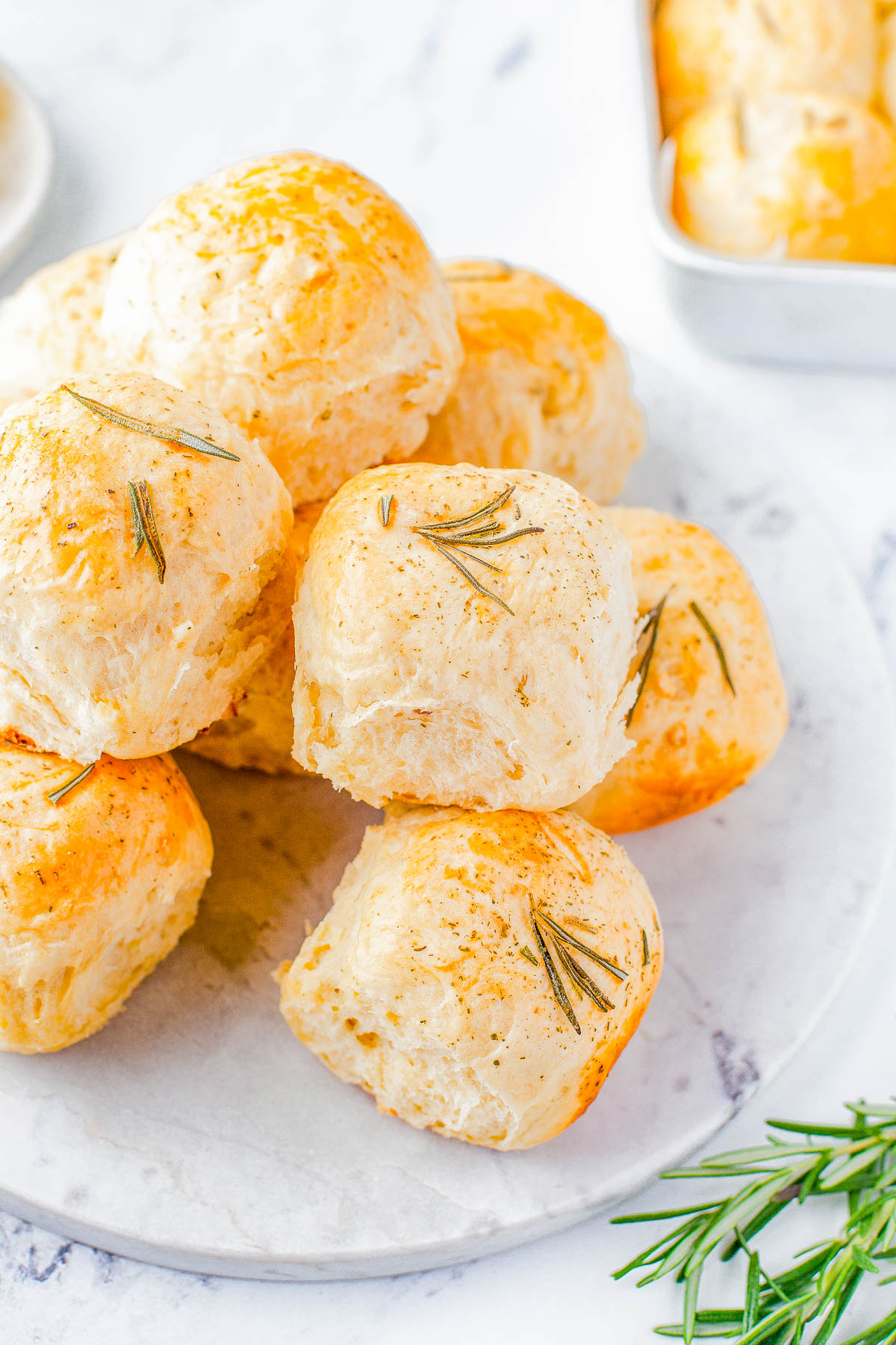 A plate of golden-brown dinner rolls garnished with fresh rosemary sprigs, with a few more rolls in a baking dish in the background.