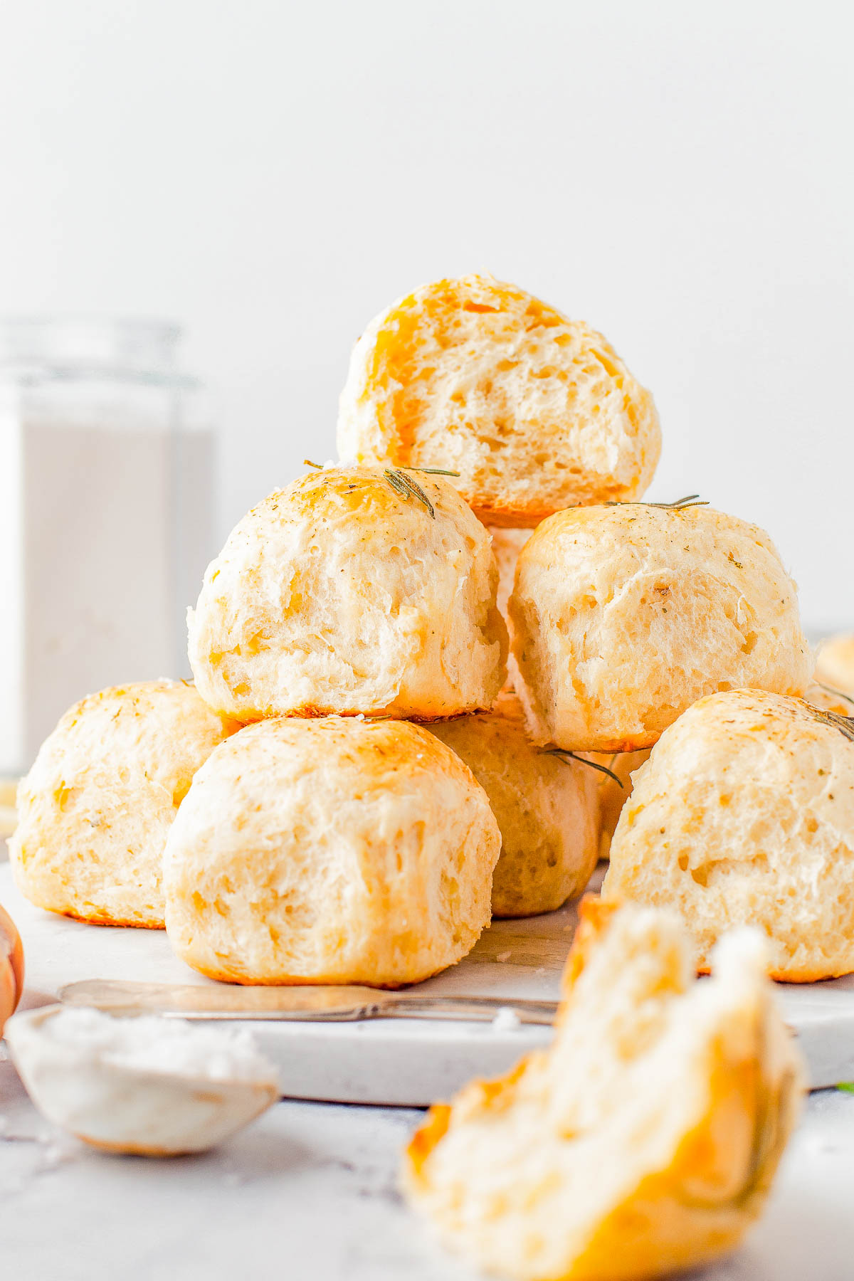 A stack of freshly baked dinner rolls on a table, with a jar of flour and a small bowl in the background. The rolls are golden-brown and fluffy.