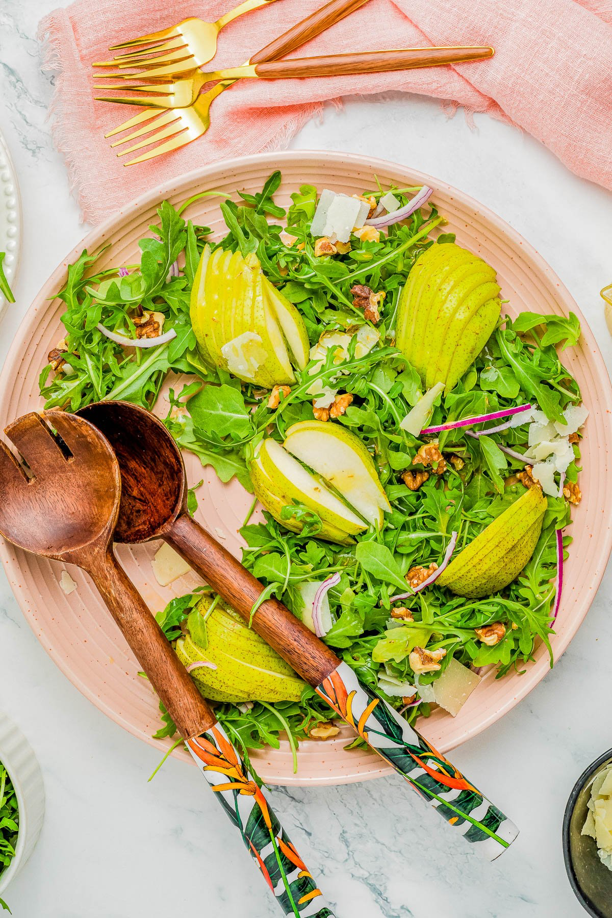 A salad of arugula, sliced pears, walnuts, and shaved cheese on a pink plate with wooden salad servers and gold utensils on a marble surface.