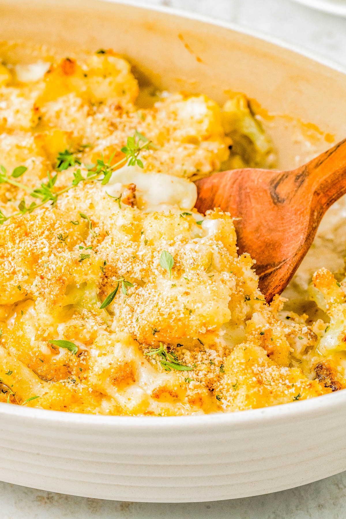 A wooden spoon scoops a portion of baked macaroni and cheese with a crispy breadcrumb topping from a white casserole dish.