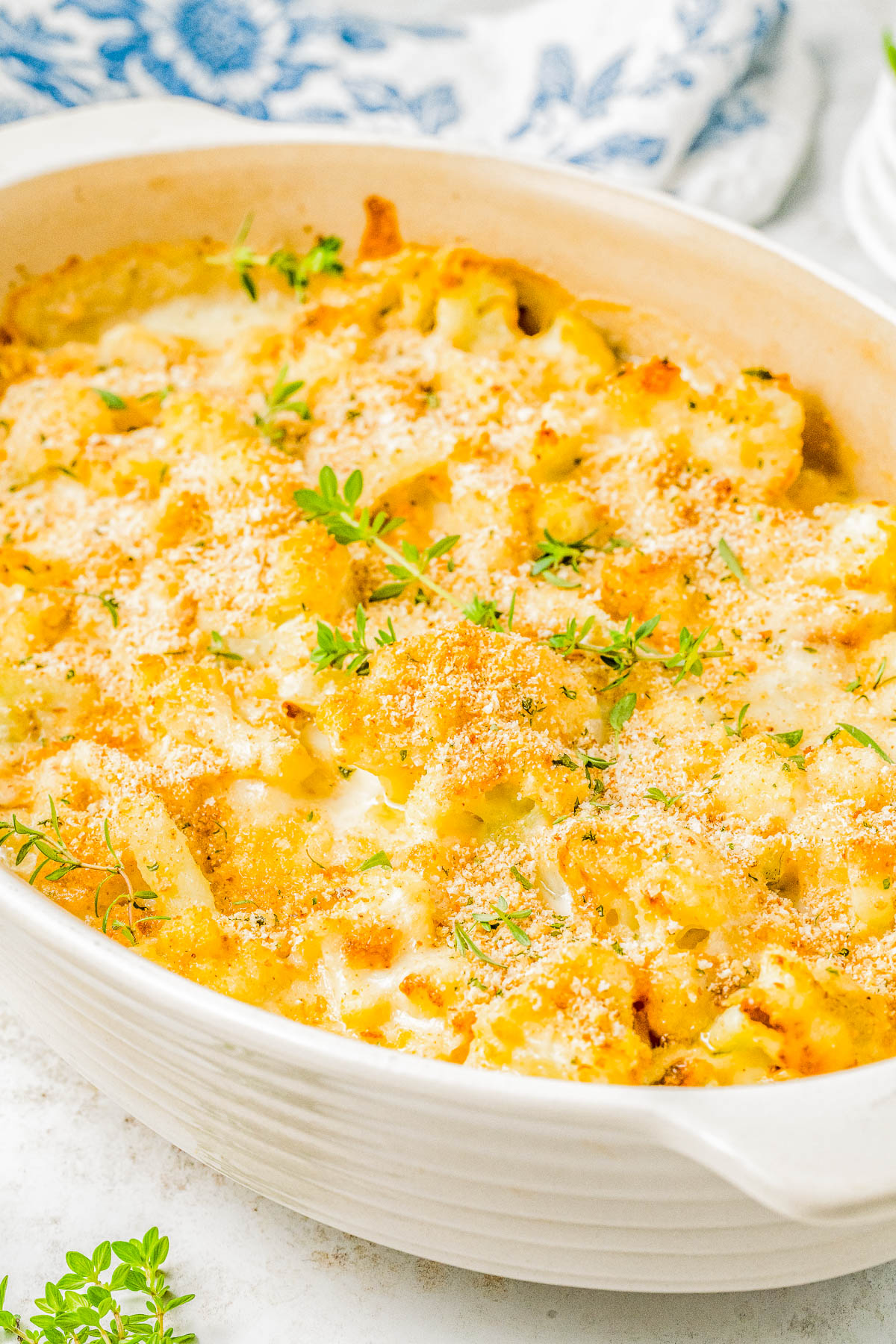 A white casserole dish filled with a baked cheesy cauliflower dish topped with breadcrumbs and garnished with fresh herbs.