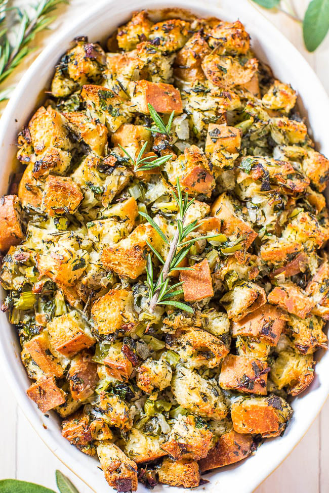 A ceramic dish filled with baked stuffing made of bread cubes, herbs, and vegetables, garnished with a sprig of rosemary.