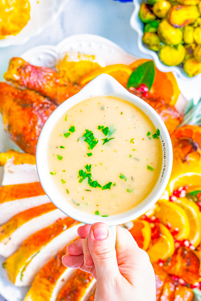 Hand holding a white gravy boat filled with creamy gravy garnished with chopped herbs, with a platter of roast turkey and assorted vegetables in the background.