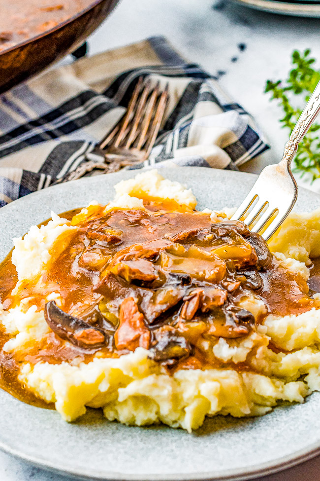 A plate of mashed potatoes topped with mushroom gravy, with a fork resting on the side, and a napkin and silverware in the background.