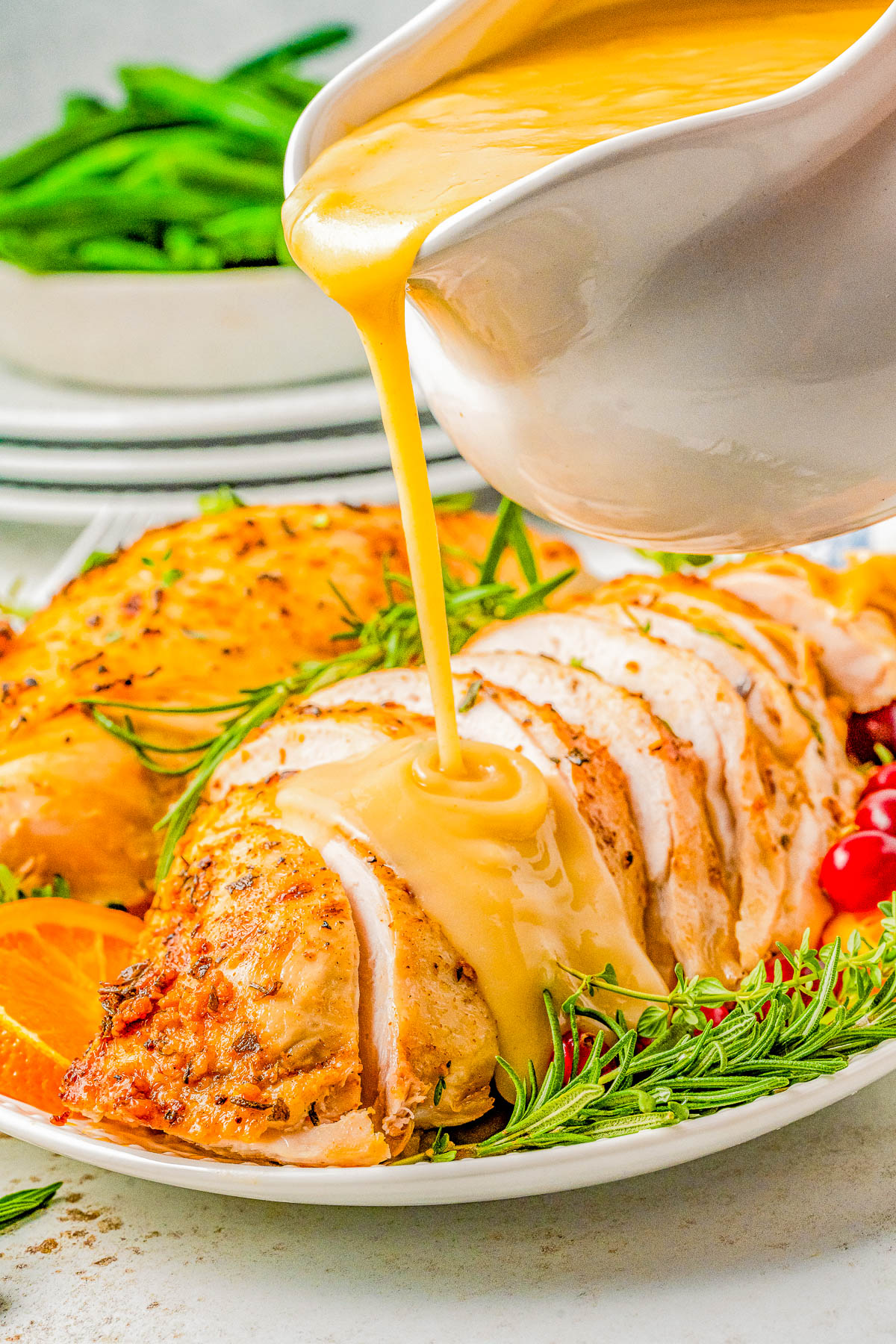 A close-up of sliced roasted turkey being drizzled with gravy from a white gravy boat. The plate is garnished with fresh rosemary and orange slices.