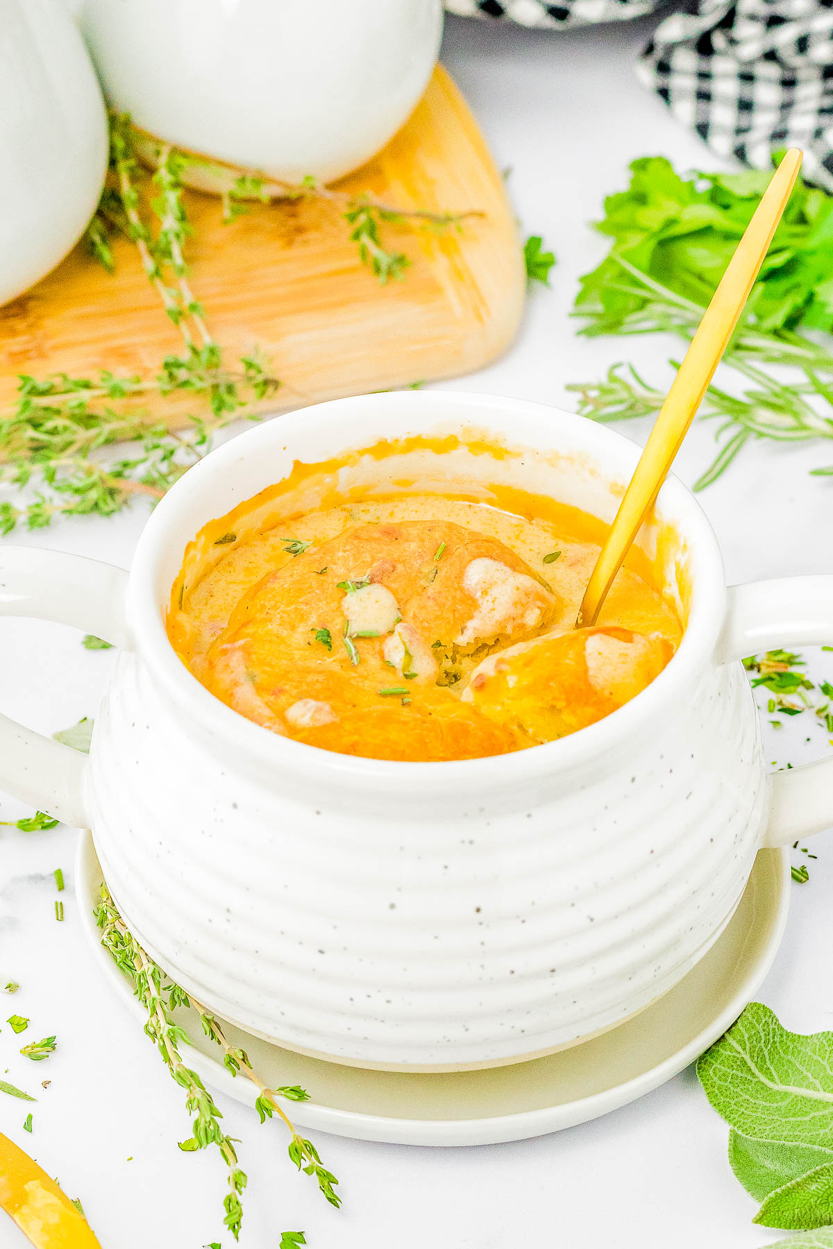 A white ceramic bowl filled with creamy pumpkin soup, garnished with fresh herbs, and a gold spoon resting inside. The bowl is placed on a white saucer with green leaves and sprigs of thyme nearby.