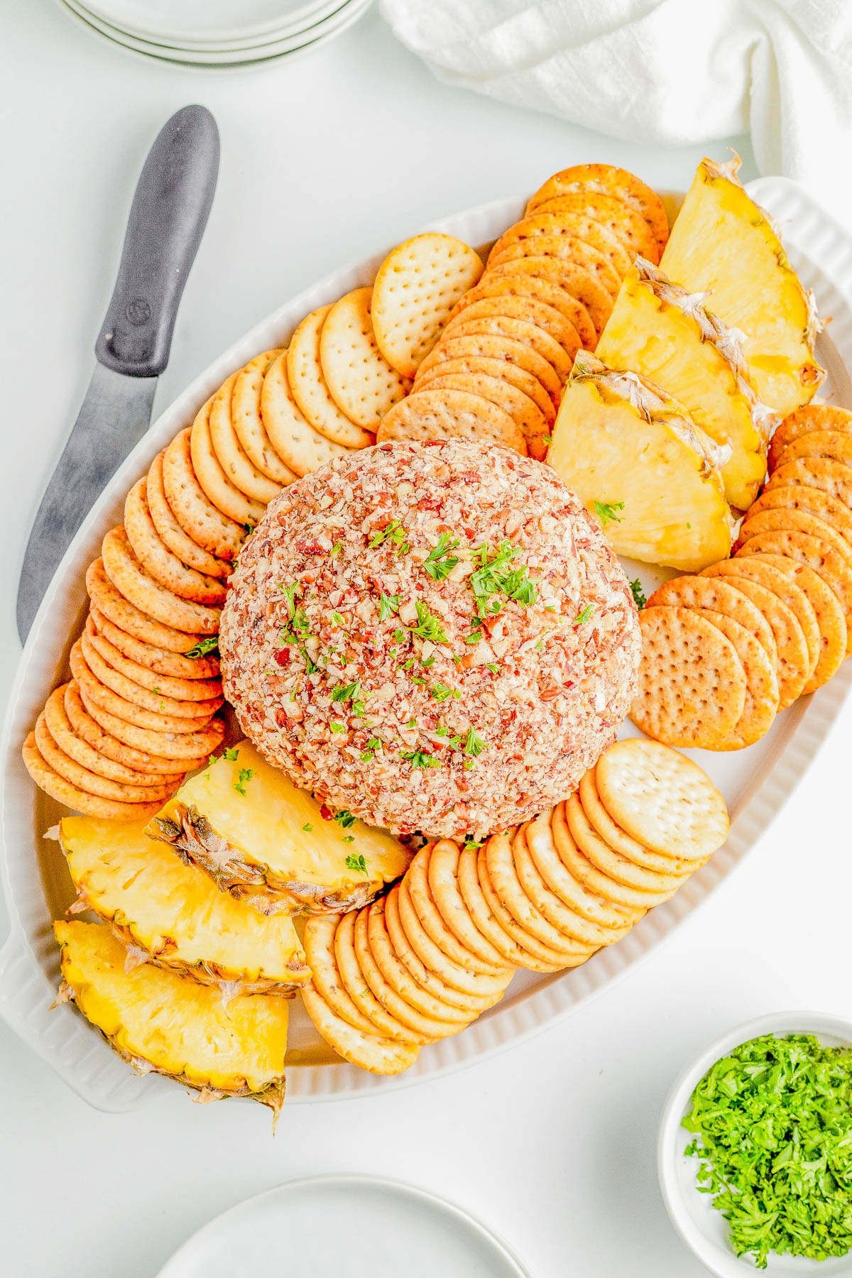 Cheese ball coated with nuts, surrounded by round crackers and pineapple slices on an oval platter, with a knife and small dish of herbs nearby.