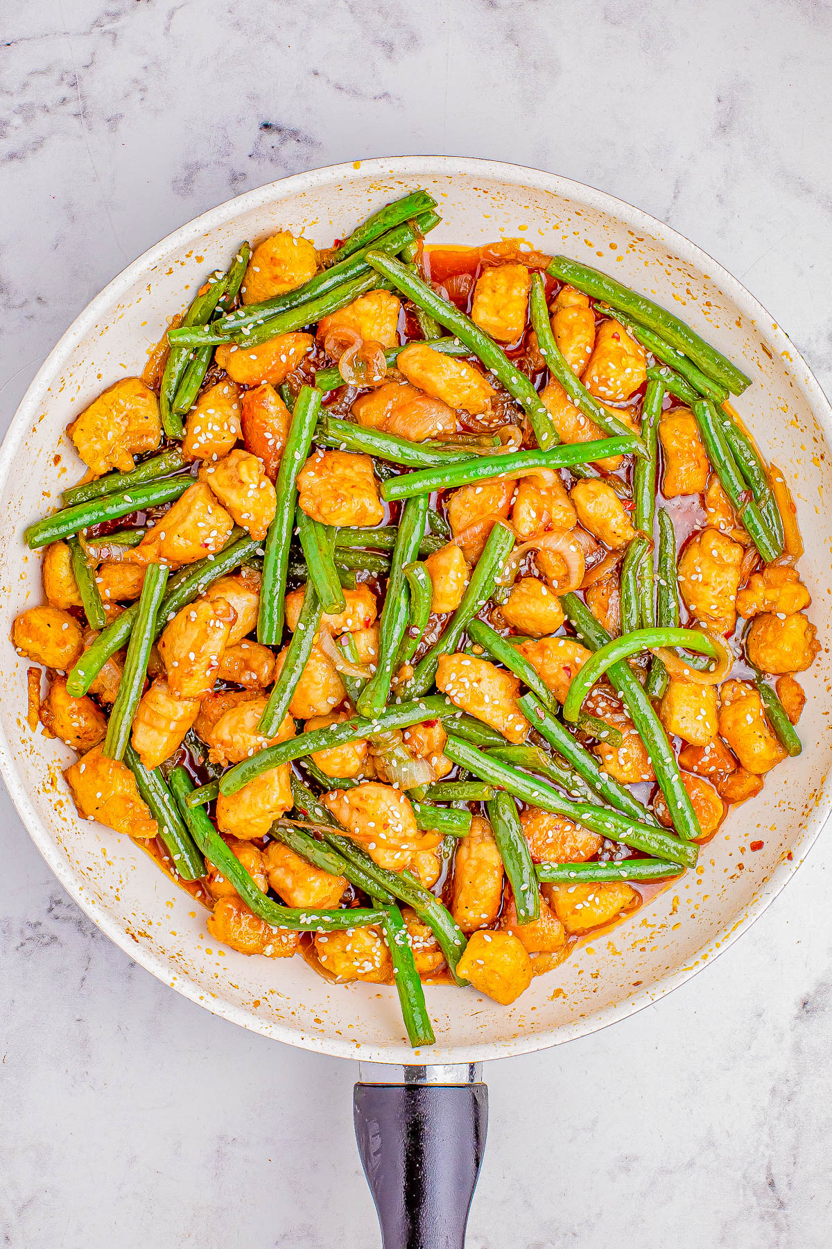 Stir-fried chicken and green beans in a sauce, served in a skillet on a marble surface.
