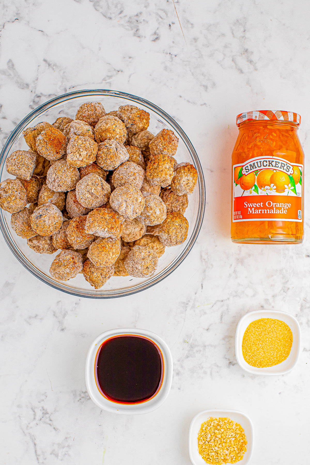 A bowl of meatballs next to a jar of smucker's sweet orange marmalade, and small bowls of seasonings.