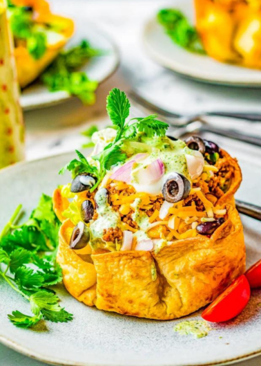 A taco salad in a crisp tortilla bowl is topped with shredded cheese, black olives, diced onions, sour cream, cilantro, and a drizzle of green sauce, set on a white plate with a red fork.