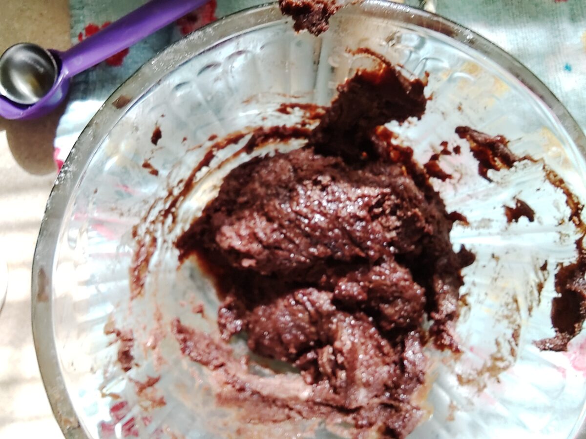 A glass bowl filled with dark brown, partially mixed batter or dough, with a small amount smeared on the side. A purple utensil is visible nearby.