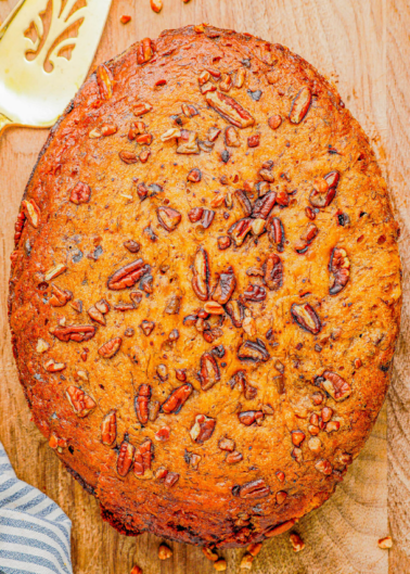 A round, baked cake with a golden-brown crust, topped with chopped pecans, placed on a wooden surface next to a brass spatula and a striped cloth.