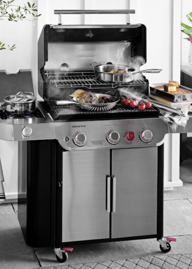 A stainless steel outdoor gas grill with various foods, including vegetables, meat, and bread, cooking on it. The grill has multiple burners, an open hood, and additional cooking accessories on the side.