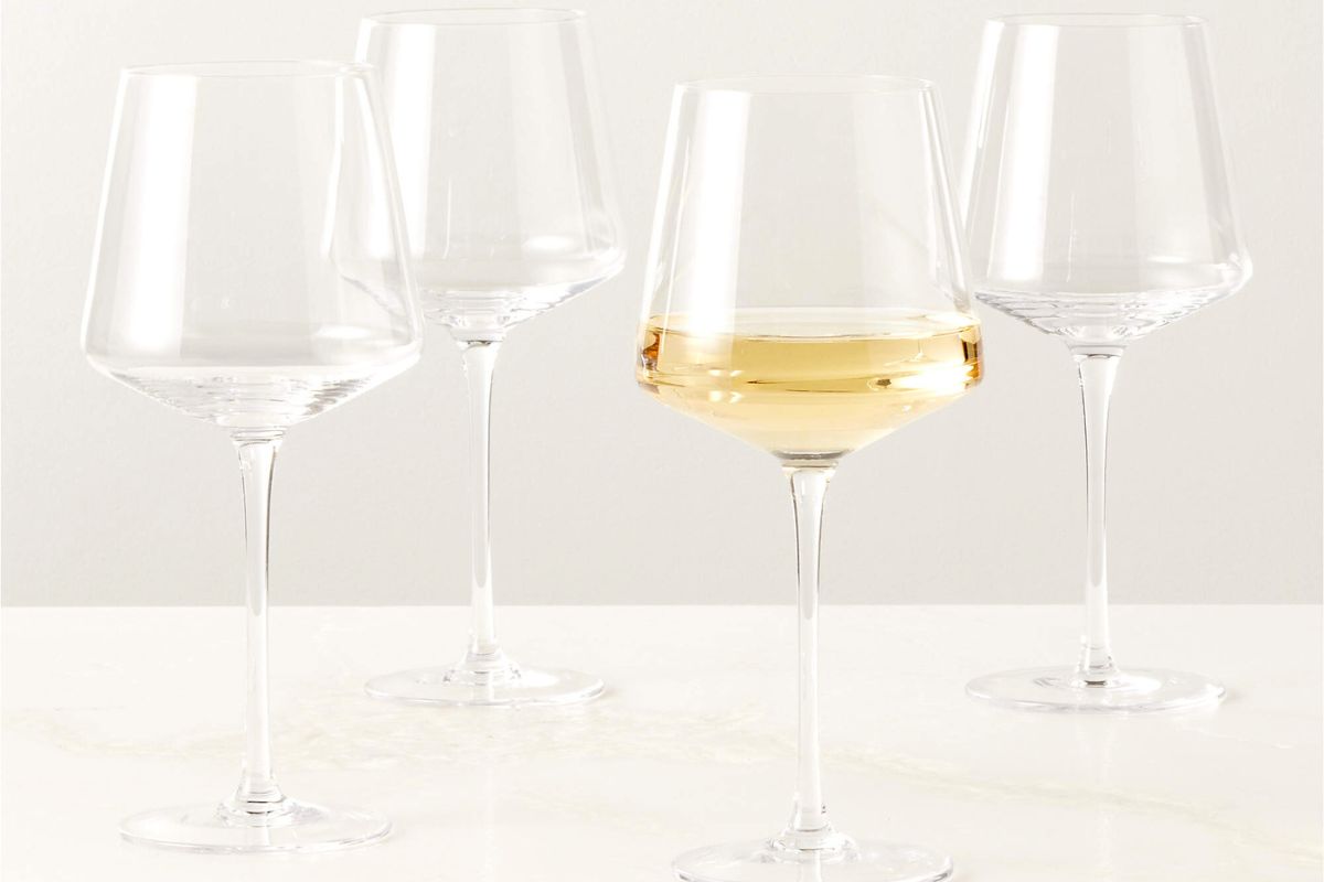 Four empty wine glasses on a white surface, with one glass partially filled with white wine.