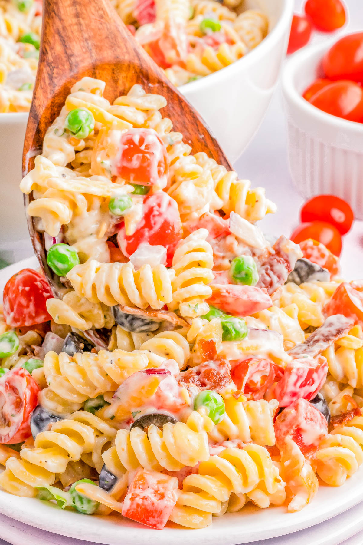 A wooden spoon scoops a portion of creamy pasta salad with rotini, cherry tomatoes, peas, and cheese from a bowl. Additional ingredients are visible in small bowls in the background.