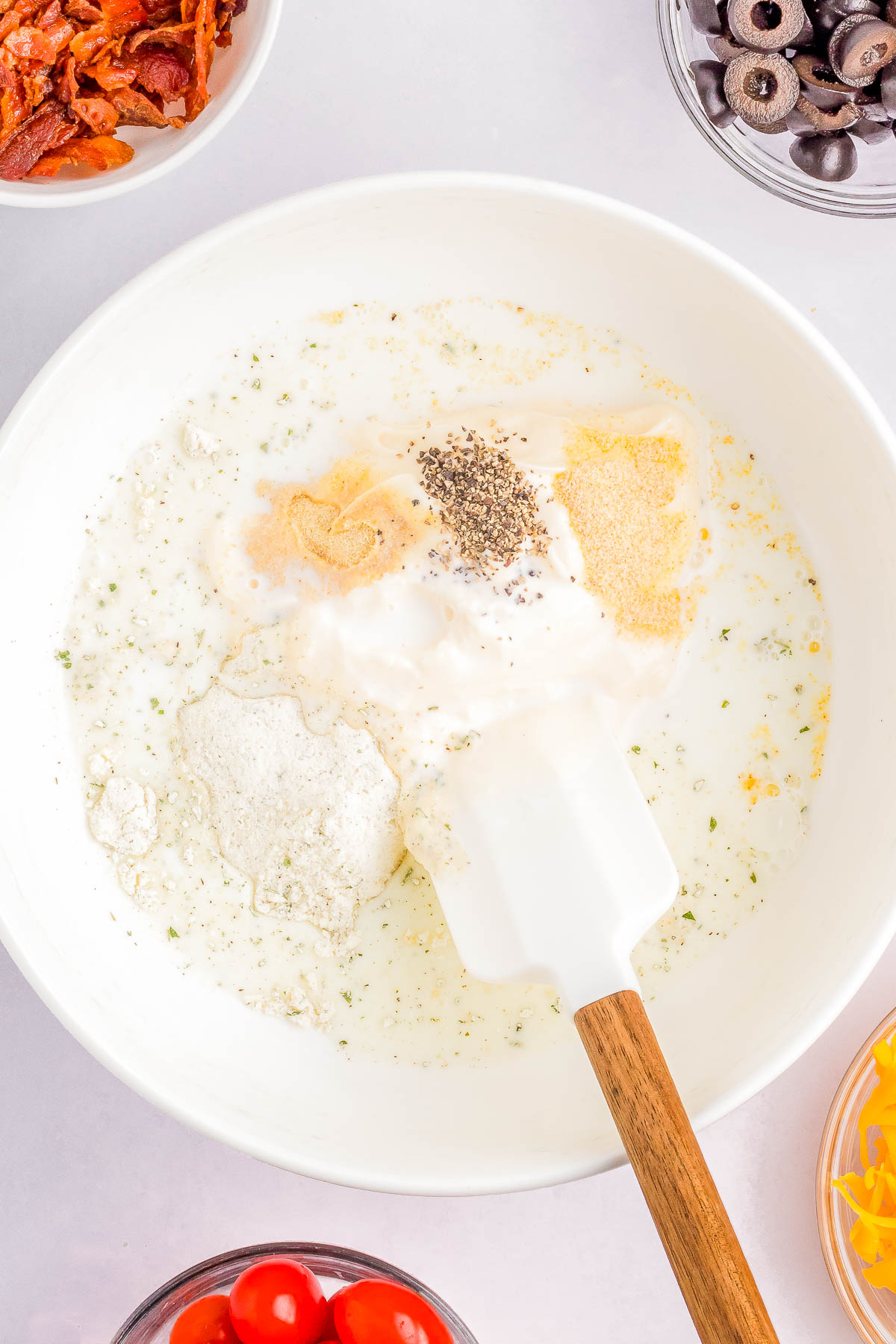 A mixing bowl contains milk, sour cream, various spices, and a spatula. Surrounding the bowl are small dishes with cherry tomatoes, grated cheese, sliced black olives, and crispy bacon bits.