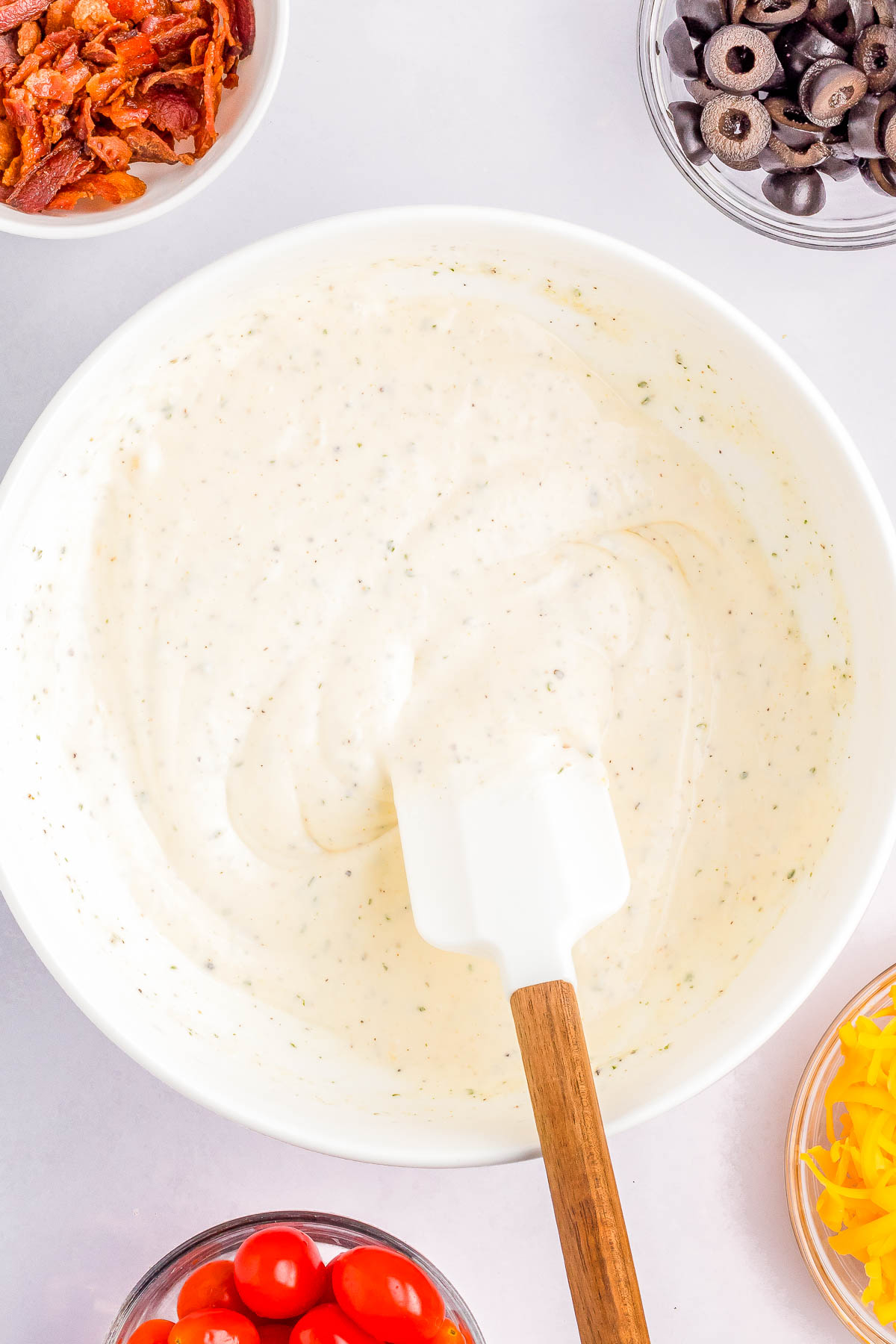 A bowl of creamy white dip with a spatula in it, surrounded by small bowls of toppings including bacon bits, black olives, grated cheese, and cherry tomatoes.