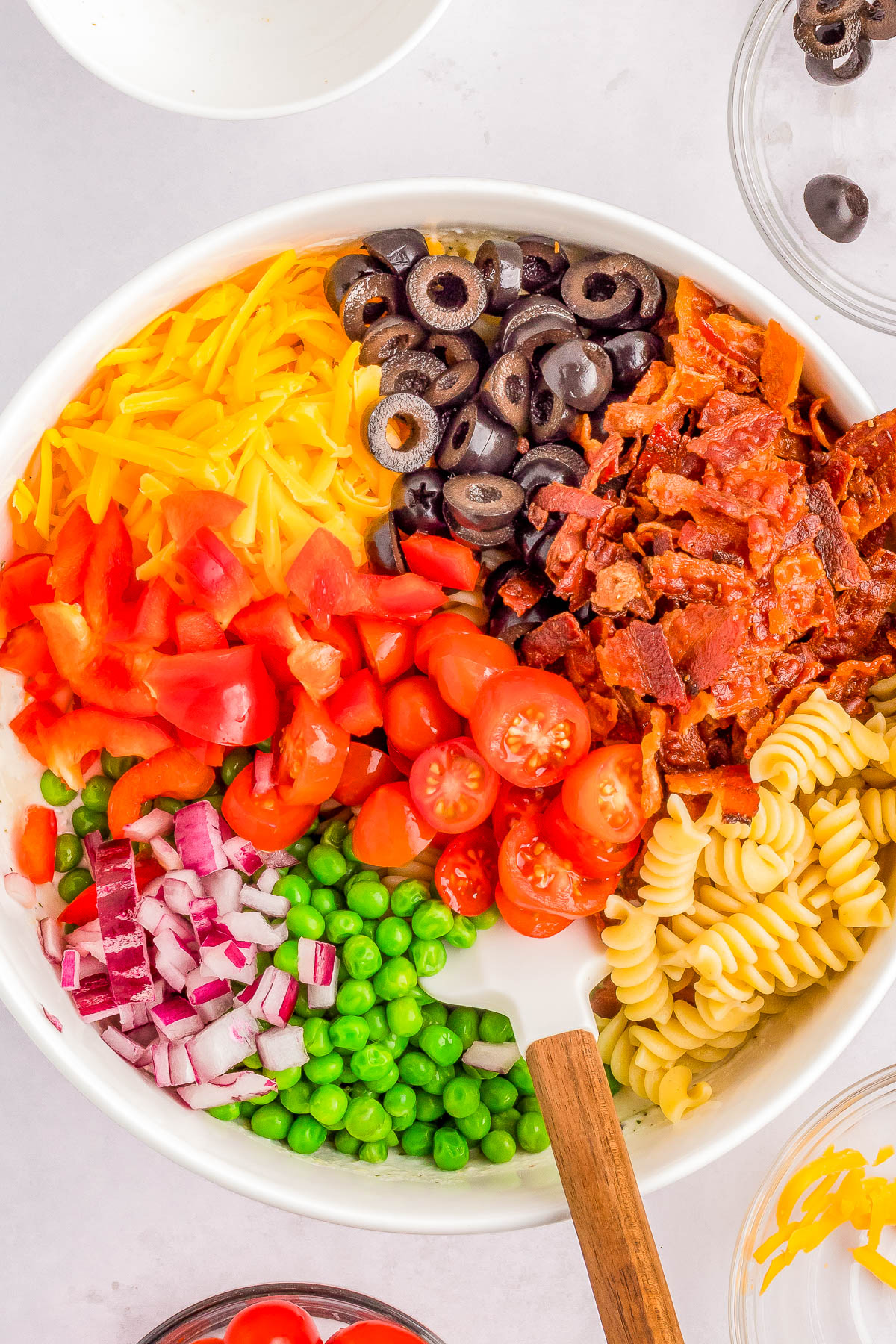 A bowl filled with rotini pasta, cherry tomatoes, black olives, crispy bacon, shredded cheddar cheese, chopped red onions, diced red bell peppers, and green peas, with a white spoon in the center.