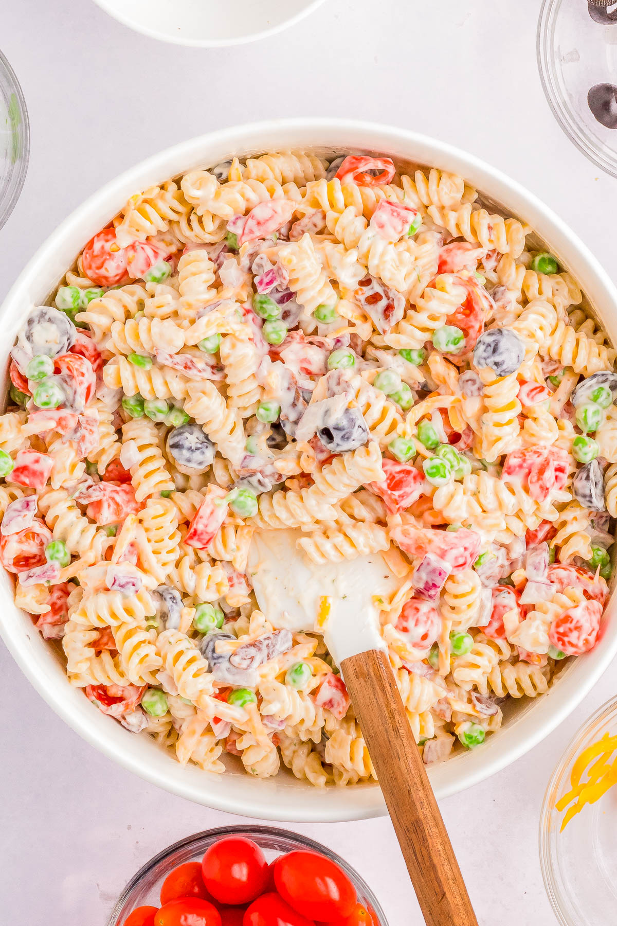 A bowl filled with creamy pasta salad containing rotini pasta, peas, cherry tomatoes, olives, and a wooden spoon for mixing.