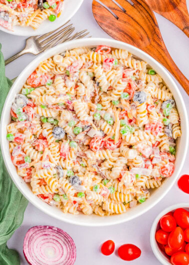 A bowl of rotini pasta salad with vegetables including peas, tomatoes, black olives, and diced red onions in a creamy dressing. Wooden serving spoons, a fork, and additional ingredients are nearby.