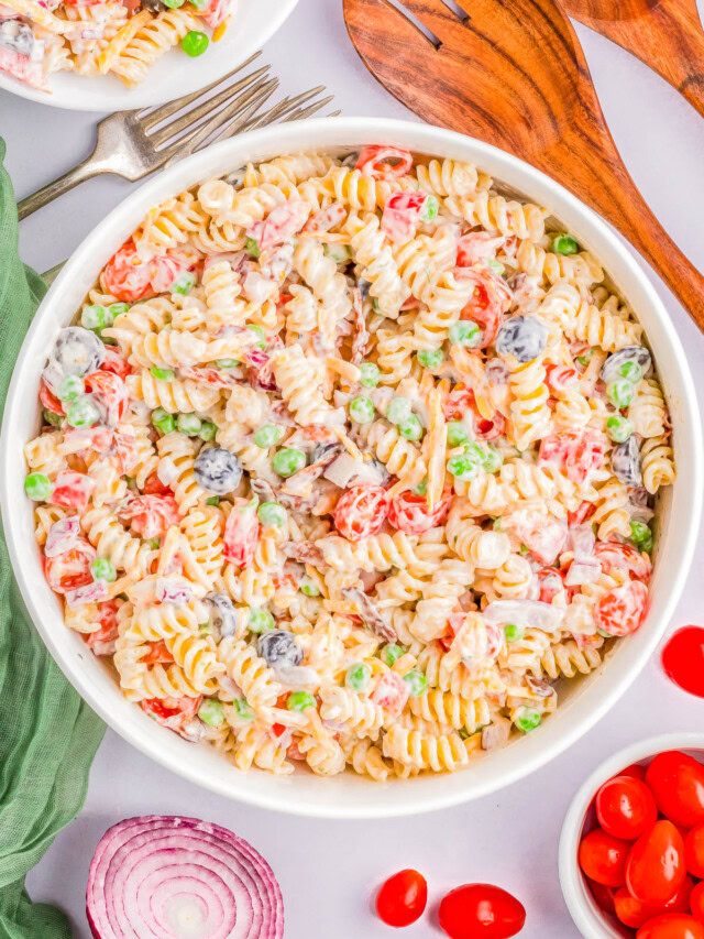 A bowl of rotini pasta salad with vegetables including peas, tomatoes, black olives, and diced red onions in a creamy dressing. Wooden serving spoons, a fork, and additional ingredients are nearby.