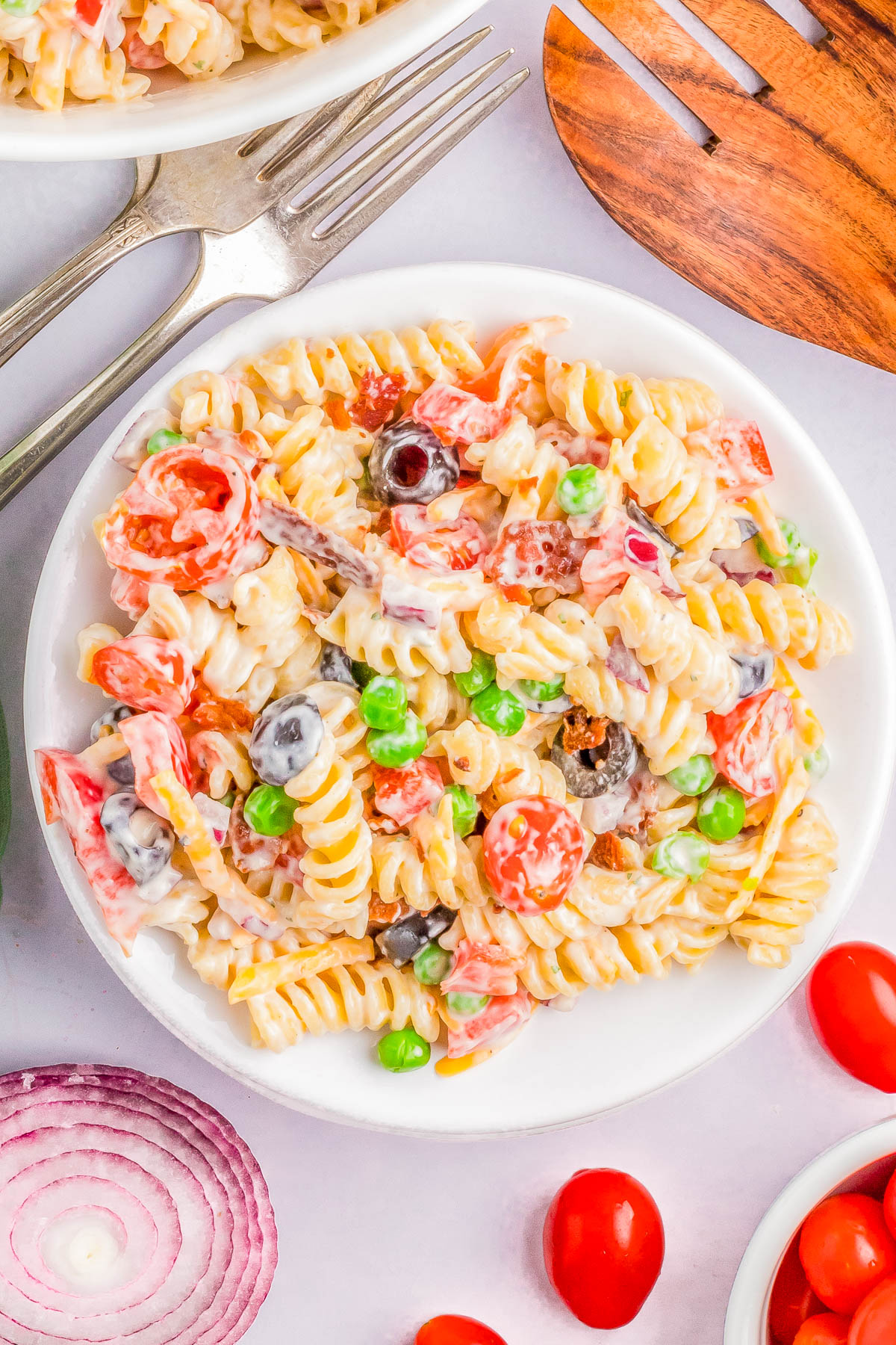 A plate of rotini pasta salad mixed with peas, cherry tomatoes, black olives, and red onion, dressed in a creamy sauce. Forks and wooden salad servers are placed nearby.