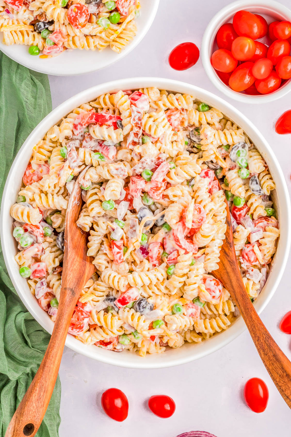 A large bowl of creamy pasta salad with rotini, peas, cherry tomatoes, and mixed vegetables. Two wooden serving spoons are placed in the bowl. Additional cherry tomatoes are near the bowl.