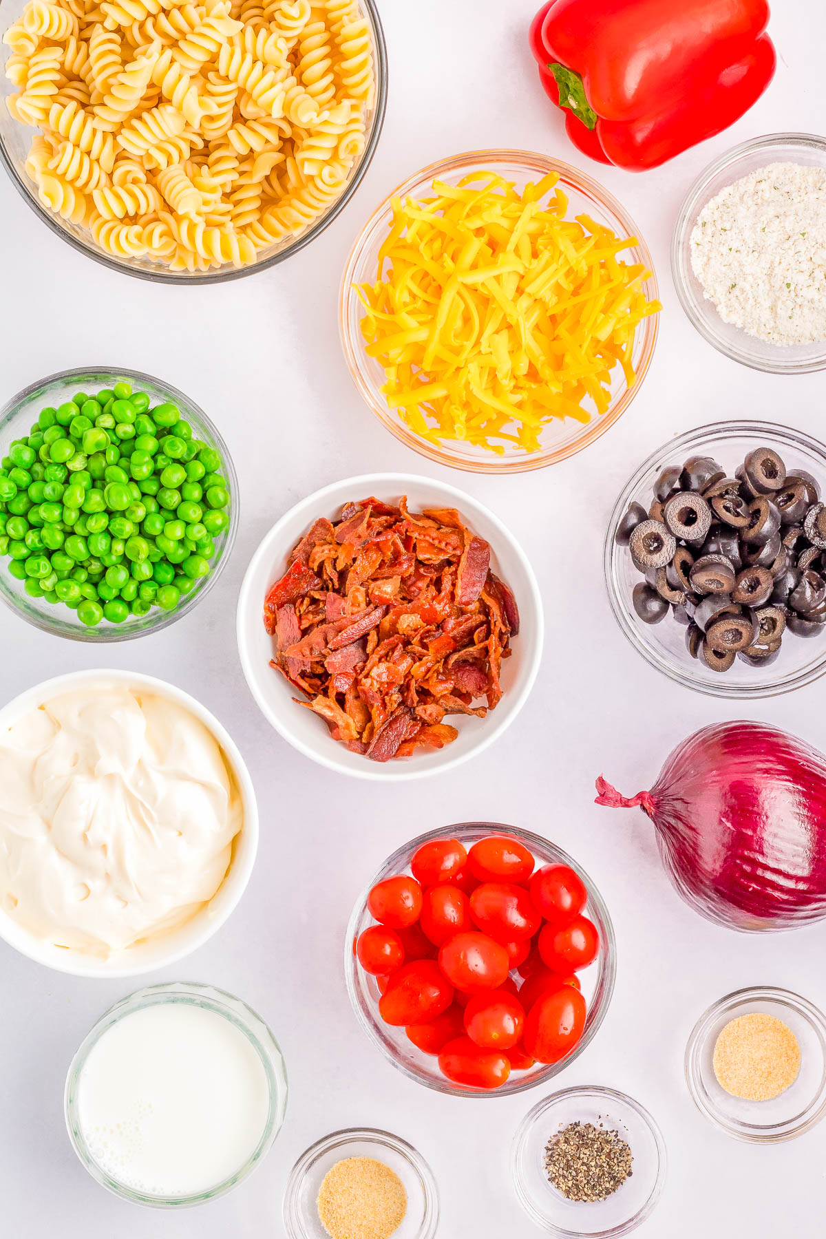 Bowls of pasta, shredded cheese, peas, bacon pieces, mayonnaise, milk, cherry tomatoes, black olives, diced red onion, red bell pepper, and various seasonings on a white surface.