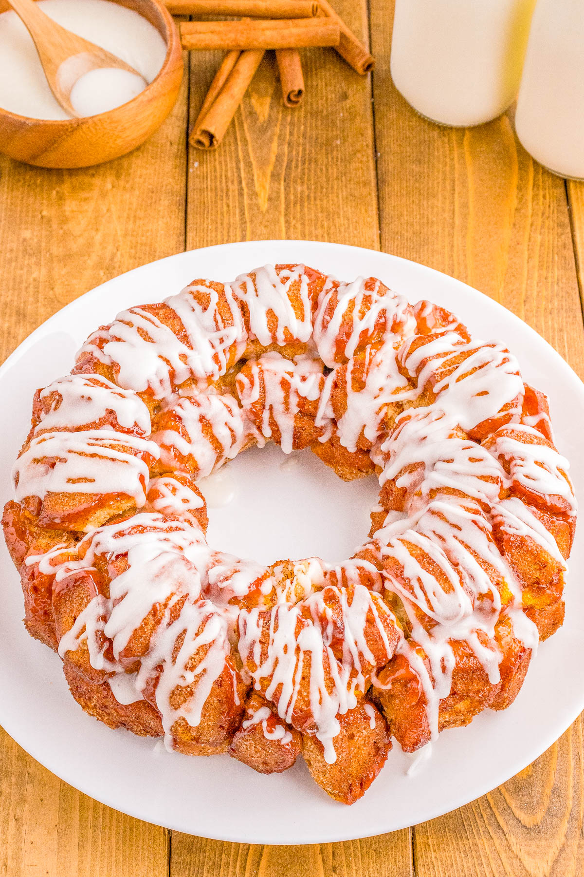 Cinnamon roll monkey bread with white icing drizzle on a white plate, surrounded by cinnamon sticks, a bowl of sugar, and a glass of milk on a wooden table.