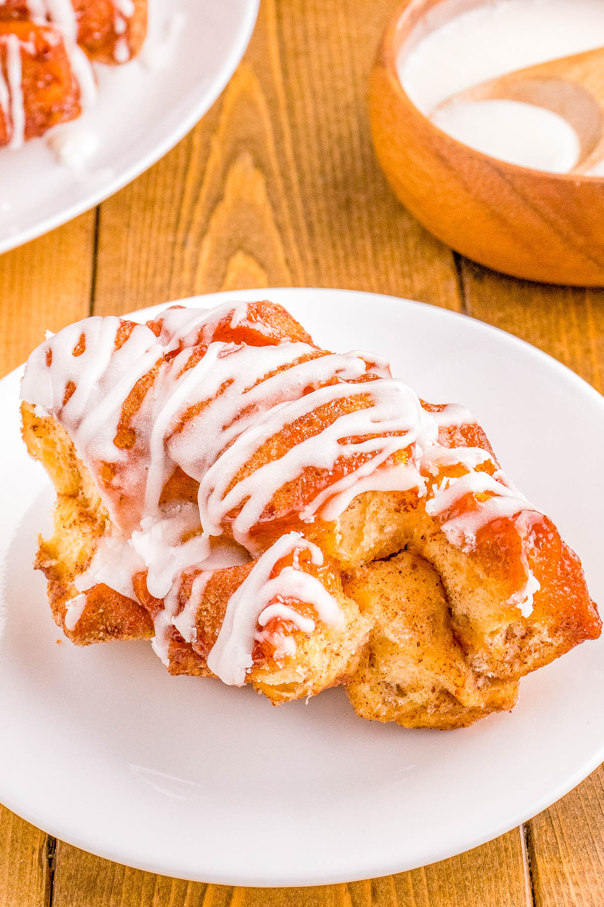 A plate containing a freshly baked cinnamon roll drizzled with white icing, on a wooden table with a bowl of extra icing in the background.