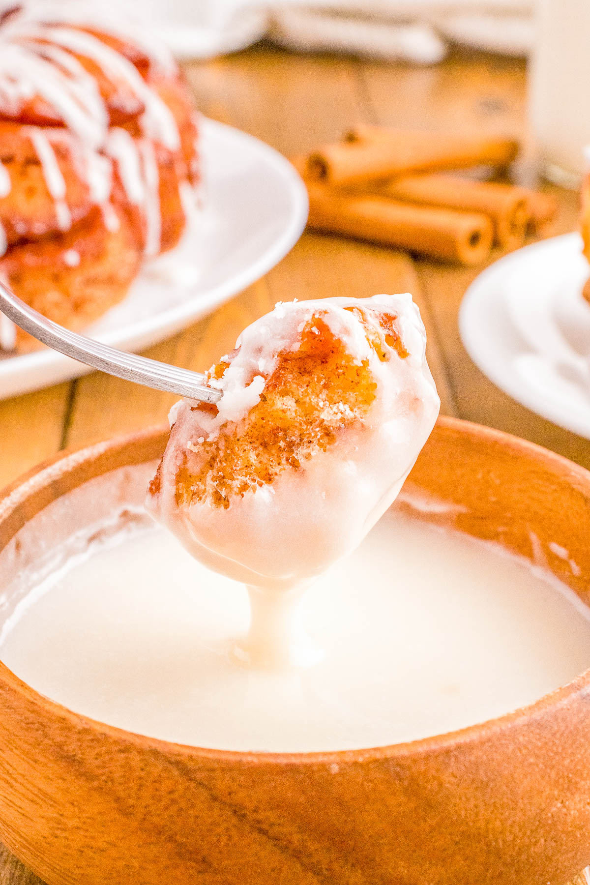A cinnamon roll being dipped into a bowl of white glaze, with additional rolls and cinnamon sticks blurred in the background.
