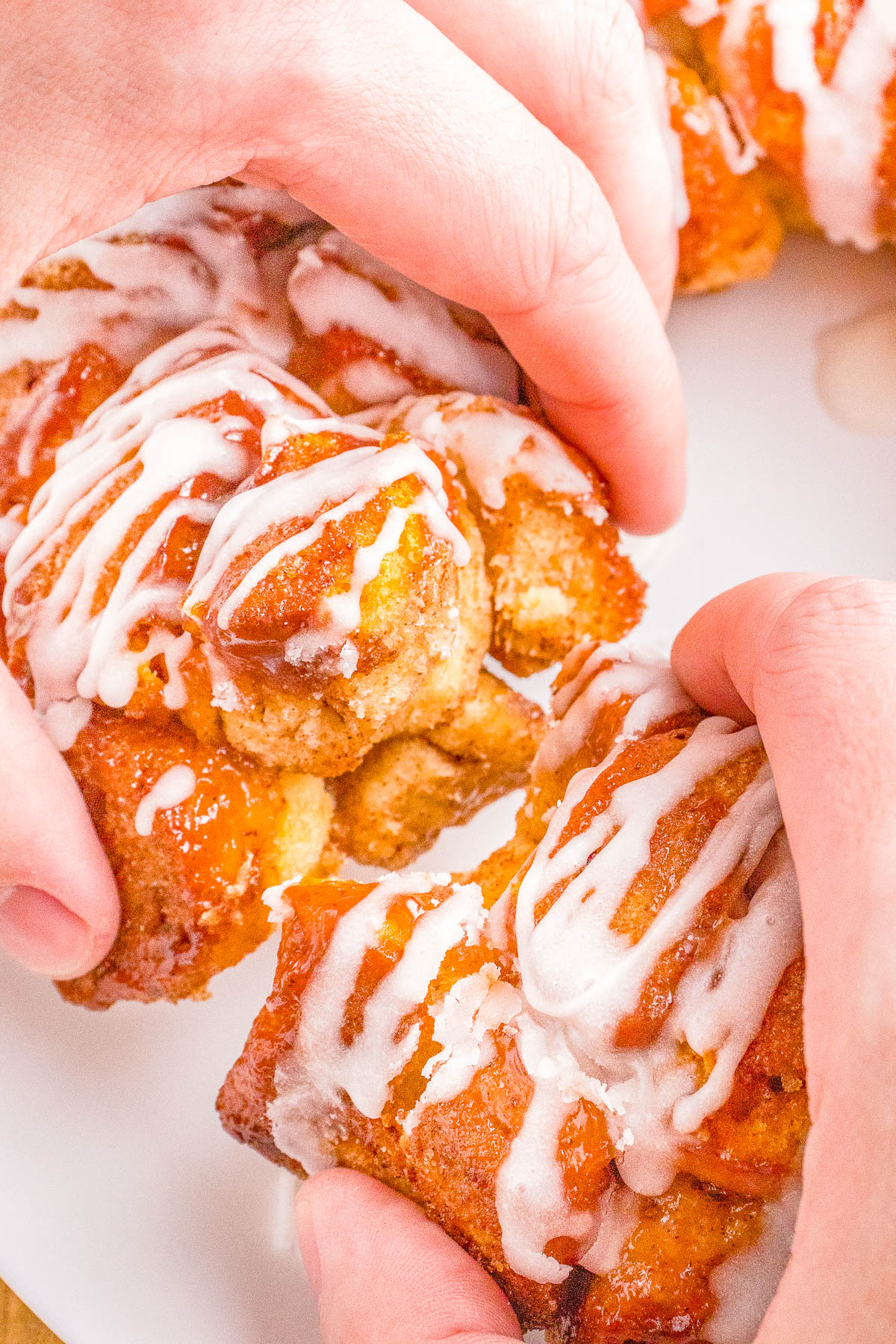 Hands holding a freshly baked cinnamon roll monkey bread covered in white icing, with more rolls visible in the background.