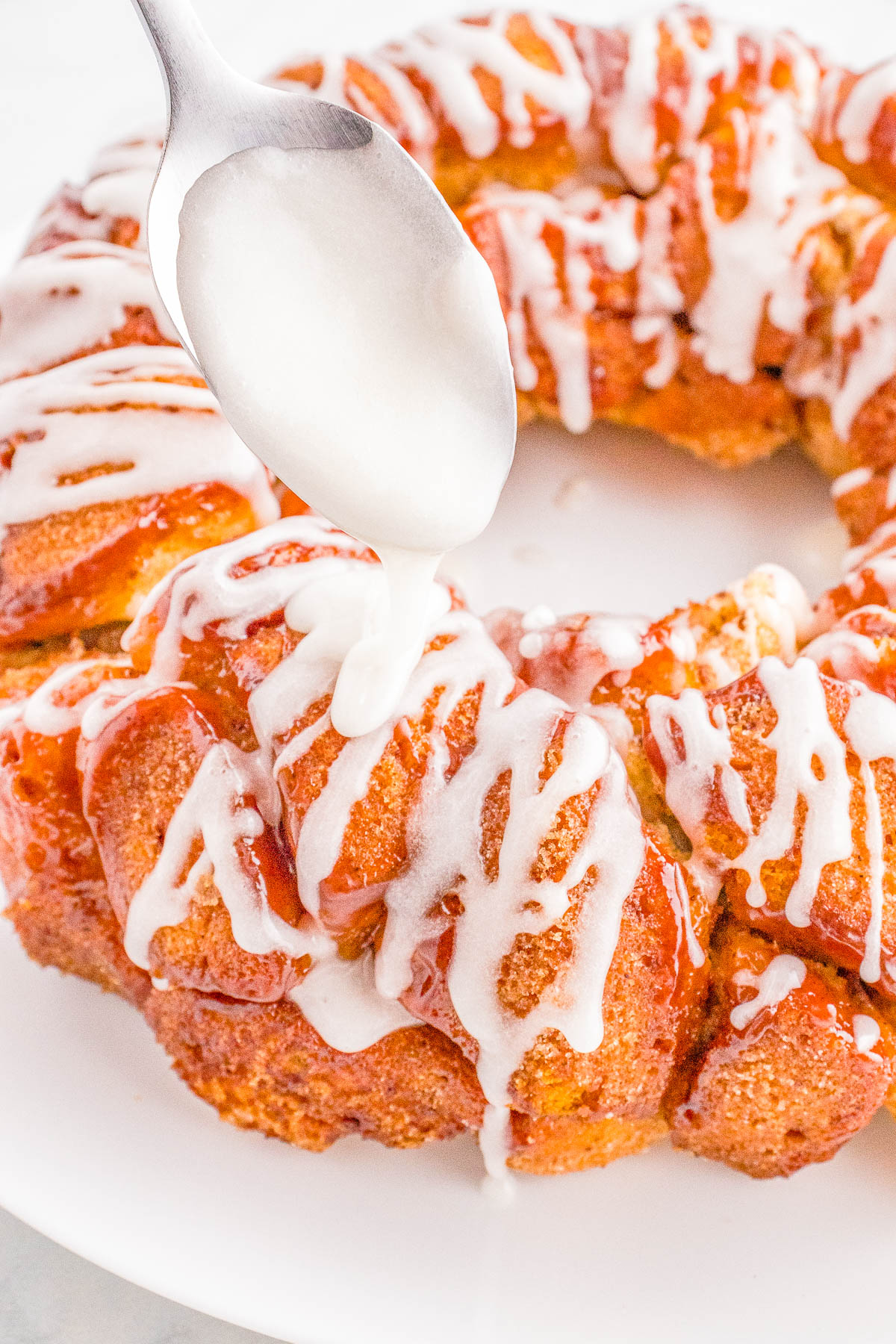 Drizzling icing on a freshly baked cinnamon roll monkey bread on a white plate.