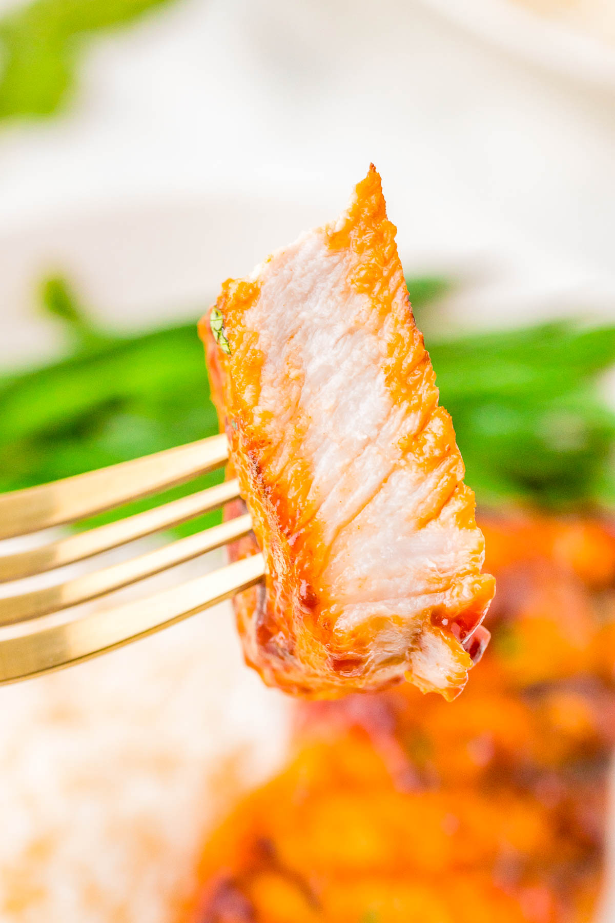 A close-up of a fork holding a piece of cooked pork with an orange glaze and white interior. Green beans are visible in the background.