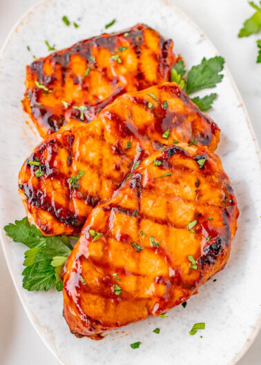 Three grilled pork chops glazed with barbecue sauce are garnished with chopped herbs and arranged on a white oval plate.