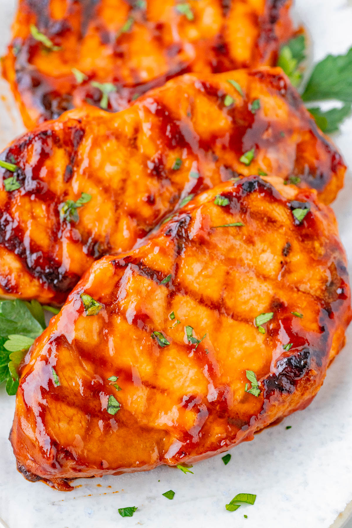 Grilled pork chops with a glossy barbecue glaze, garnished with chopped parsley, served on a white plate with a side of green herbs.