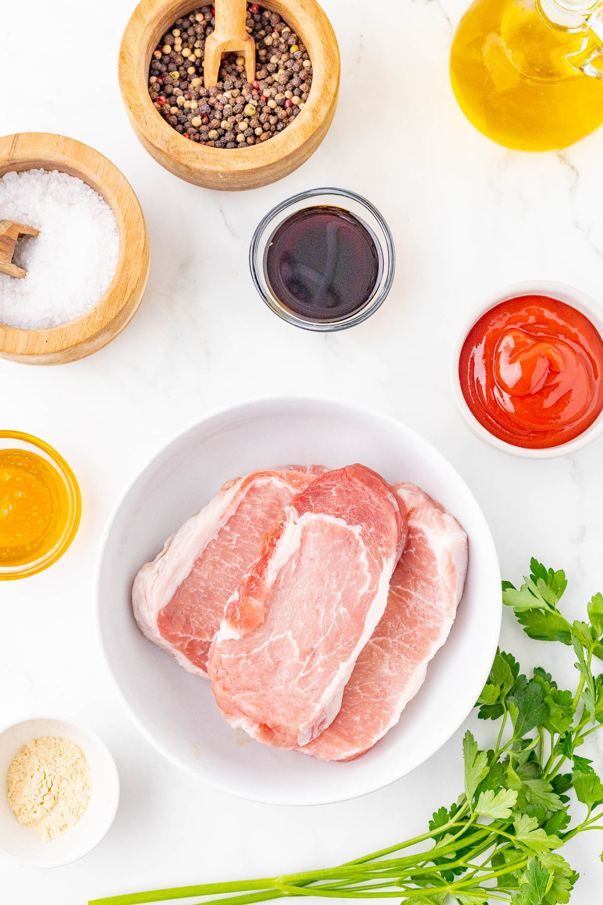 Raw pork chops in a bowl surrounded by seasonings and condiments including salt, peppercorns, soy sauce, ketchup, honey, garlic powder, and a bottle of oil on a marble countertop.